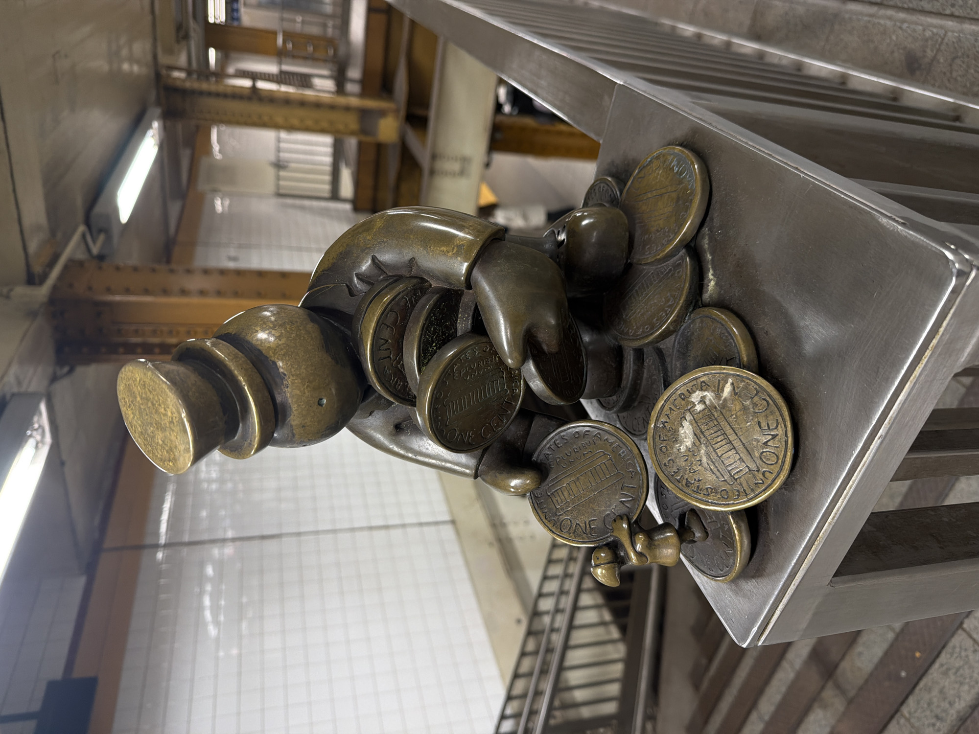 The image depicts a bronze sculpture of a hand clutching pennies, installed on a metal railing. The pennies are U.S. one-cent coins featuring the Lincoln Memorial. The sculpture is situated within a New York City subway station, as evidenced by the tiled walls, exposed structural steel beams, and staircase visible in the background.