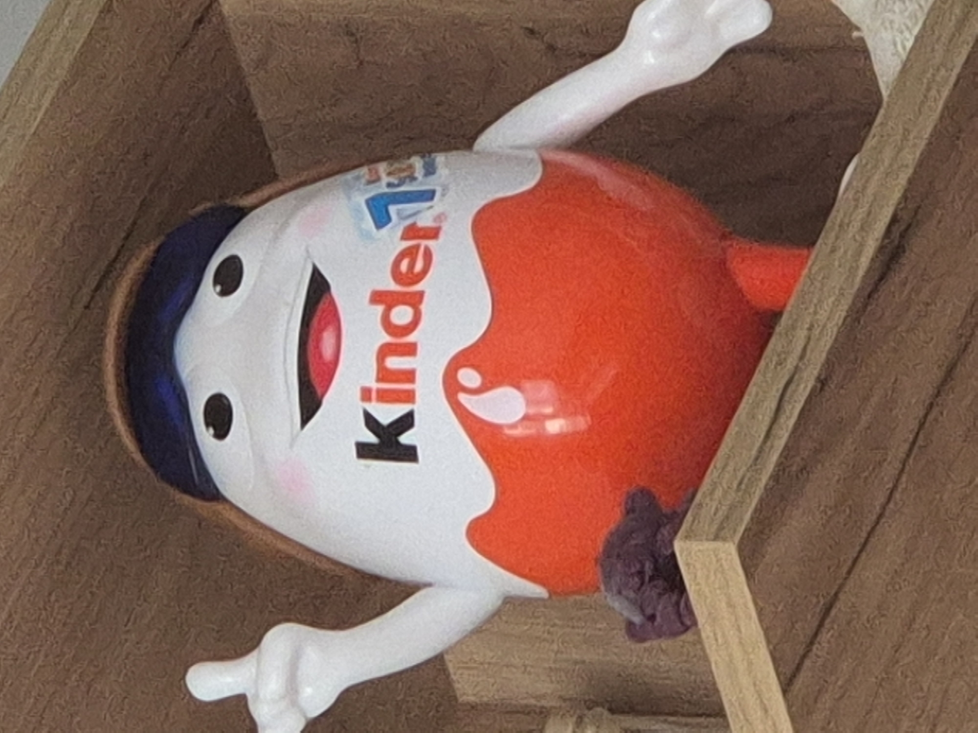 A close-up, angled shot shows a Kinder Surprise egg toy with its arms and legs extended. The toy has a white face with a red mouth, black eyes, and rosy cheeks. It wears a blue cap and a white wrapper with the "Kinder" logo prominently displayed in black and red lettering. A "1" is also visible on the wrapper. The toy is situated within a wooden structure, possibly a shelf or box, with textured brown wood forming the background and edges. A sliver of lighter wood is visible on the right side, running diagonally. A small, dark purple, fluffy object is attached to the toy's body near its right arm. The lighting appears to be indoor, with no specific indication of the time of day or weather. The overall scene suggests a playful or decorative arrangement of the toy within its wooden enclosure.