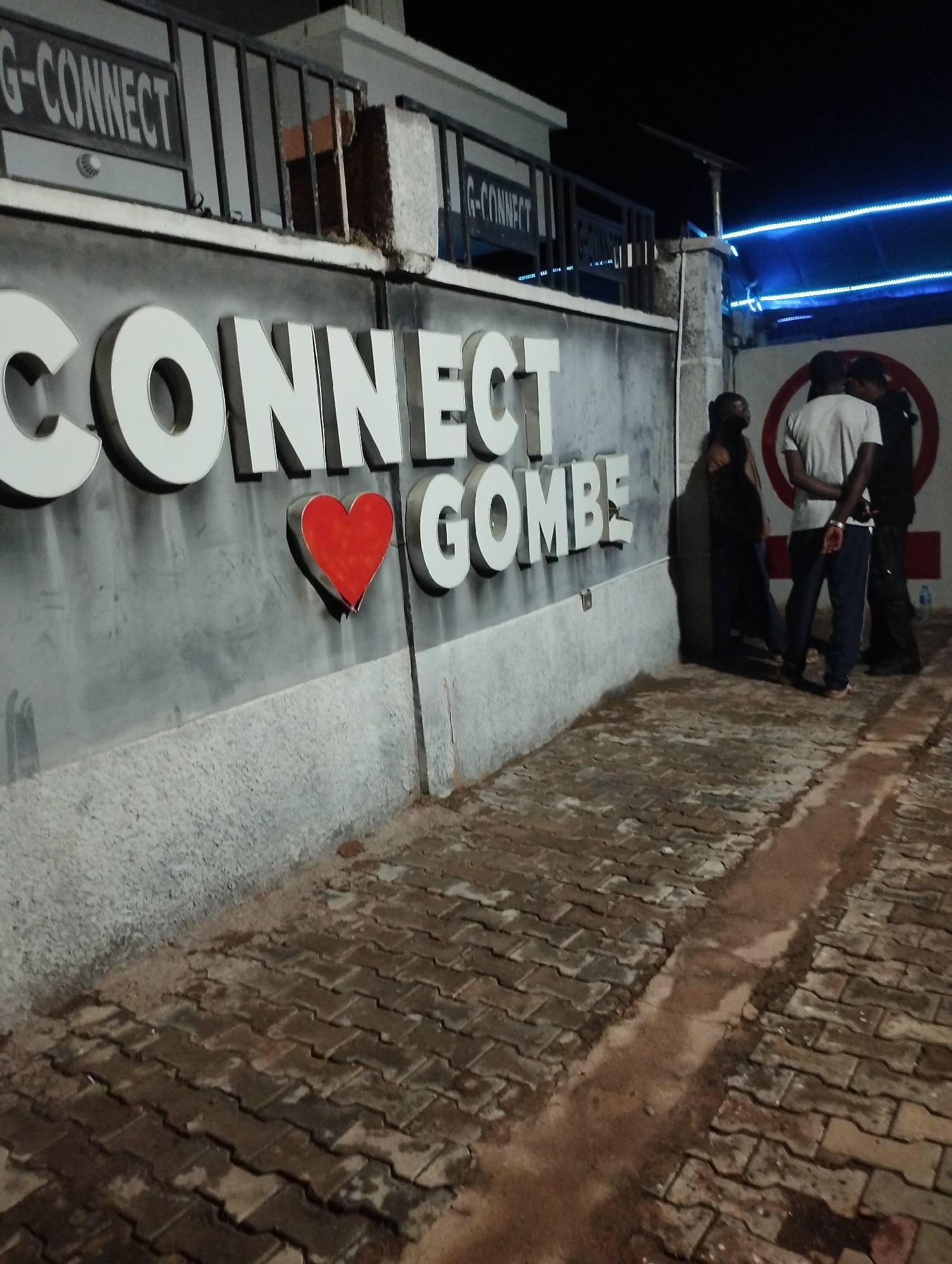 The scene shows an outdoor night shot in Gombe, Nigeria, specifically at "G-Connect Gombe." The building facade has the word "CONNECT" in white block letters, with a red heart symbol replacing the letter "O." The surface of the facade is weathered and textured. A partially obscured sign above reads "G-CONNECT."
Three men are positioned near the building's corner. One man is in a light-colored t-shirt and jeans. A second man is partially visible. A third man is closer to the building corner. Their specific interactions and activities are not clear.
The ground is paved with brown, rectangular paving stones separated by narrow channels. Some areas show dampness or discoloration. Overhead, blue LED lights are visible.