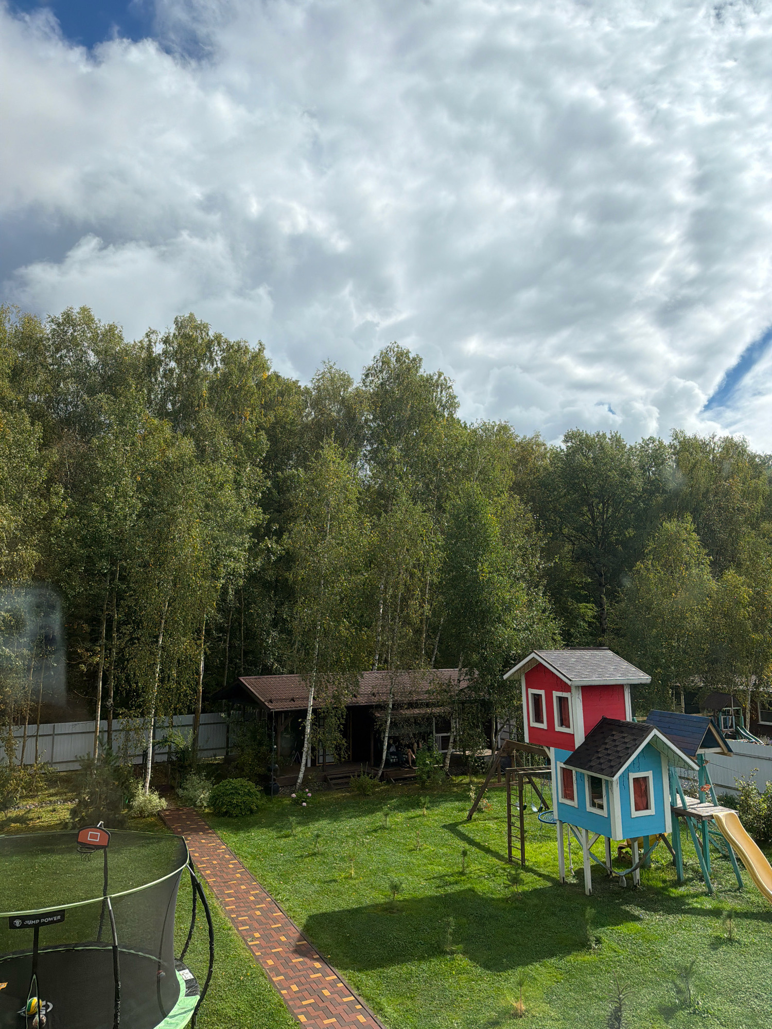 The image shows a backyard in a suburban or rural setting, possibly in Stupino, Russia, given the context. The sky is partly cloudy with large white and gray clouds. In the foreground, a trampoline with a basketball hoop is visible on the left side. A brick pathway leads from the foreground towards a wooded area.
The middle ground features a lawn with a playground. The playground includes two small, elevated playhouses, one red and one blue, connected by a swing set and a slide. Behind the playground, there is a covered outdoor seating area or gazebo with a brown roof. Further back, a fence runs along the perimeter of the property, with a dense line of trees behind it, forming a forest. The trees are a mix of deciduous and possibly some birch trees, with lush green foliage indicating a late summer or early autumn season. There is no clear indication of any specific activities or people engaged in actions, though some figures may be faintly visible within the covered structure. The overall scene depicts a recreational outdoor space adjacent to a natural wooded area.