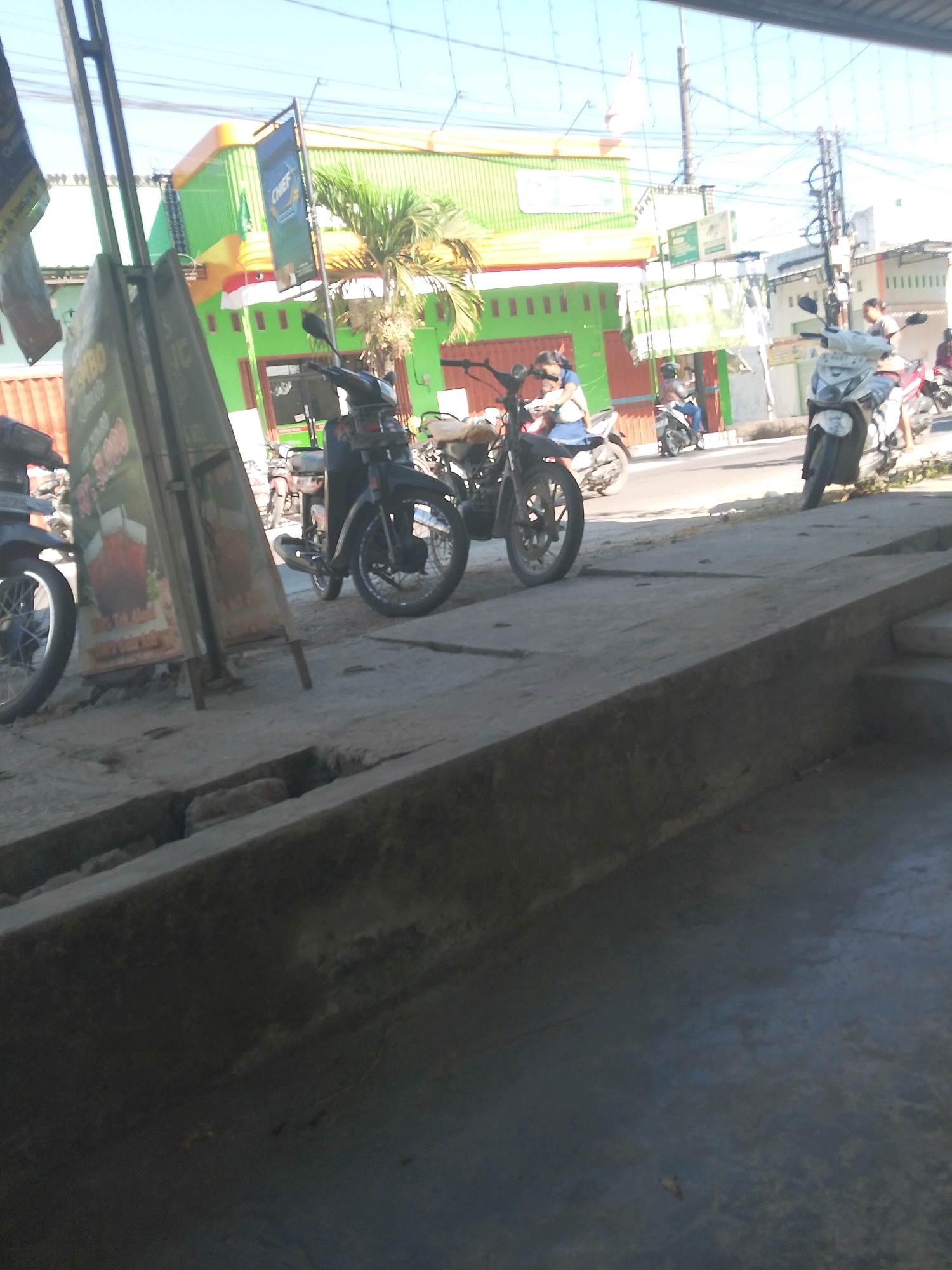 In Jampet, Indonesia, a street scene unfolds under a clear sky. The foreground features a concrete barrier casting shadows, with a narrow drainage ditch visible. Beyond this, a sidewalk hosts several parked motorcycles, their tires resting on the ground. A prominent signpost stands to the left, bearing what appears to be advertising content. In the mid-ground, a vibrant green building with orange trim dominates the background, adorned with a red and white Indonesian flag. Several people are present, some on motorcycles, others standing nearby, engaging in typical street activities. The presence of various vehicles and a commercial building suggests a bustling area.