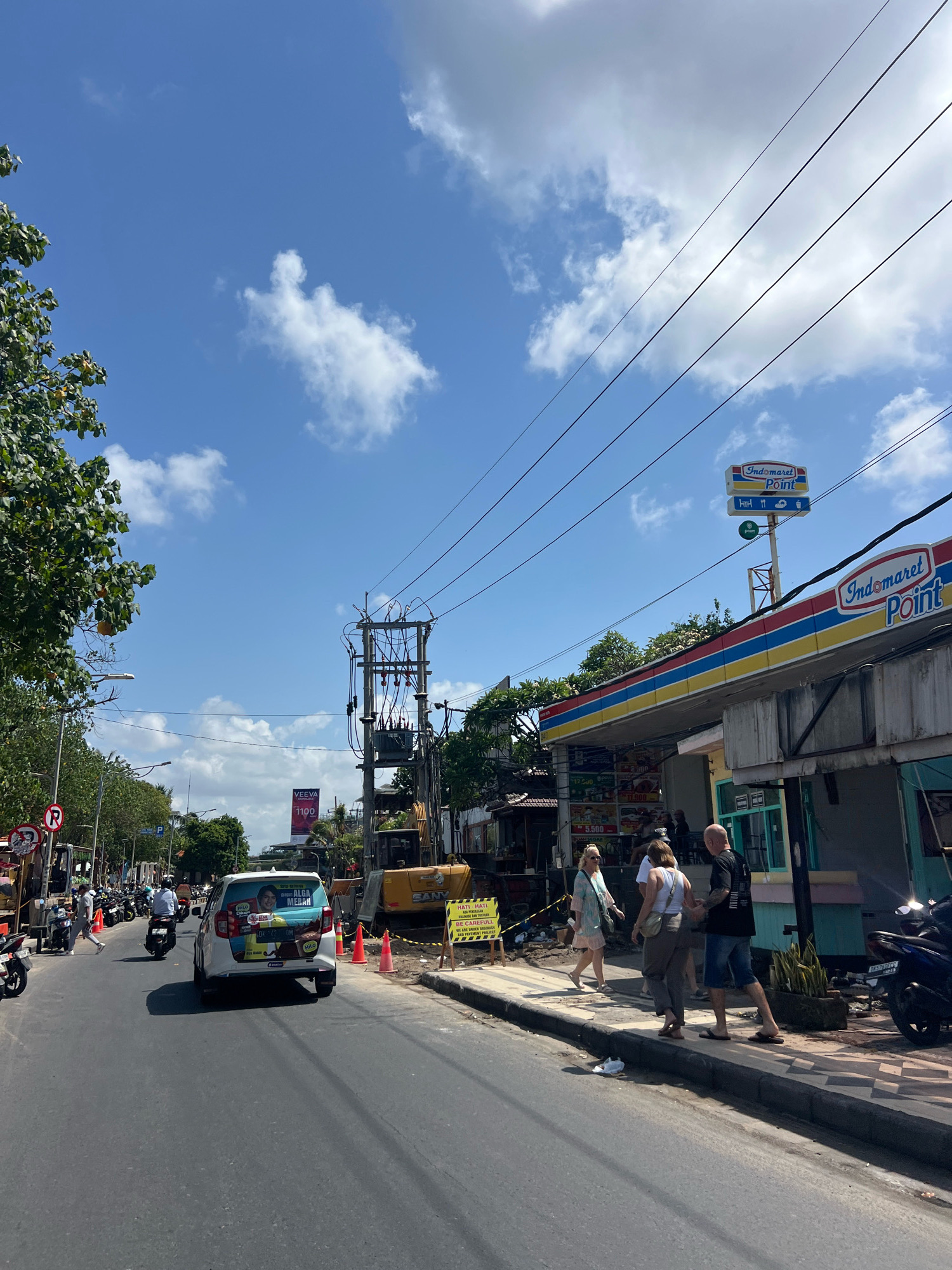 The image shows a street scene in Kuta, Indonesia, on a bright, sunny day with a blue sky and scattered white clouds. The foreground is dominated by a paved road with light traffic, including cars and numerous motorbikes. On the right side of the road, there is a brightly colored Indomaret Point convenience store with a blue, yellow, and red striped awning. A prominent sign for "Indomaret Point" is visible above the store. Several people are walking on the sidewalk in front of the store. On the left side of the road, there are trees, a traffic light, and more motorbikes parked. Further down the street, a utility pole with a large transformer and numerous wires is visible. Construction equipment, including a yellow excavator, is present on the side of the road, suggesting ongoing work or repairs. A large billboard advertising "VEEVA 1100" is also visible in the distance. The overall atmosphere is one of a busy, active commercial area in a tropical climate.