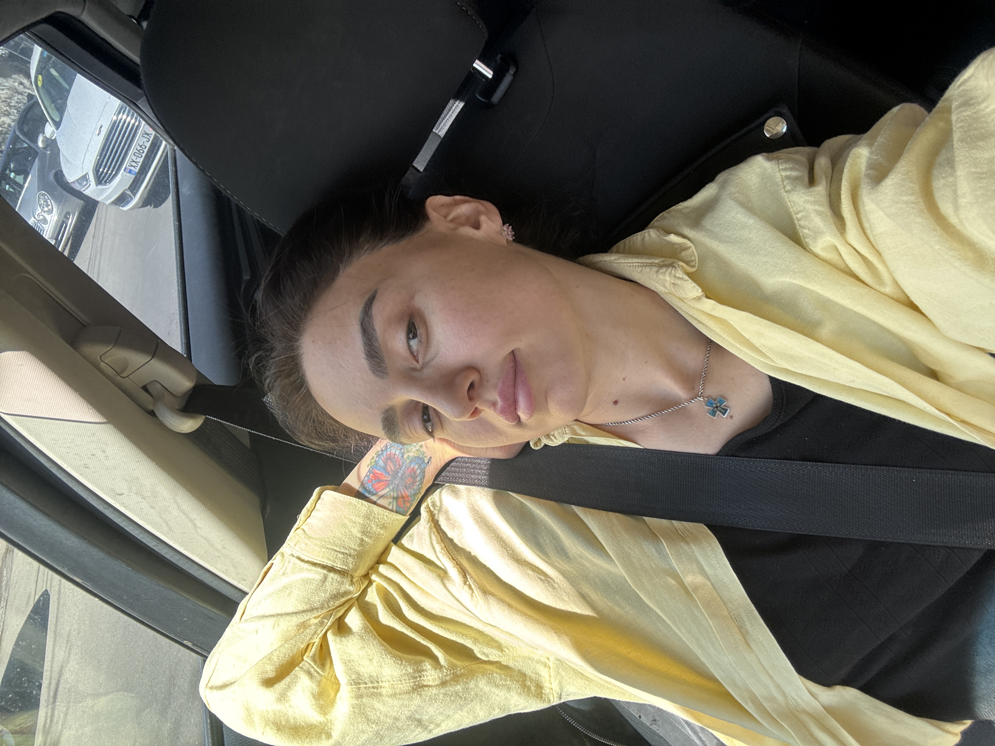 A woman is in a car. She is wearing a yellow shirt over a black top. A black seatbelt is over her. She has dark hair, and fair skin, and a butterfly tattoo on her left wrist. The car's interior is black and grey. Through the front windshield, part of a silver car and a white car are visible, the white car's license plate reads "XX-066-JX". The scene is in Tbilisi, Georgia, evidenced by the vehicle license plate code.