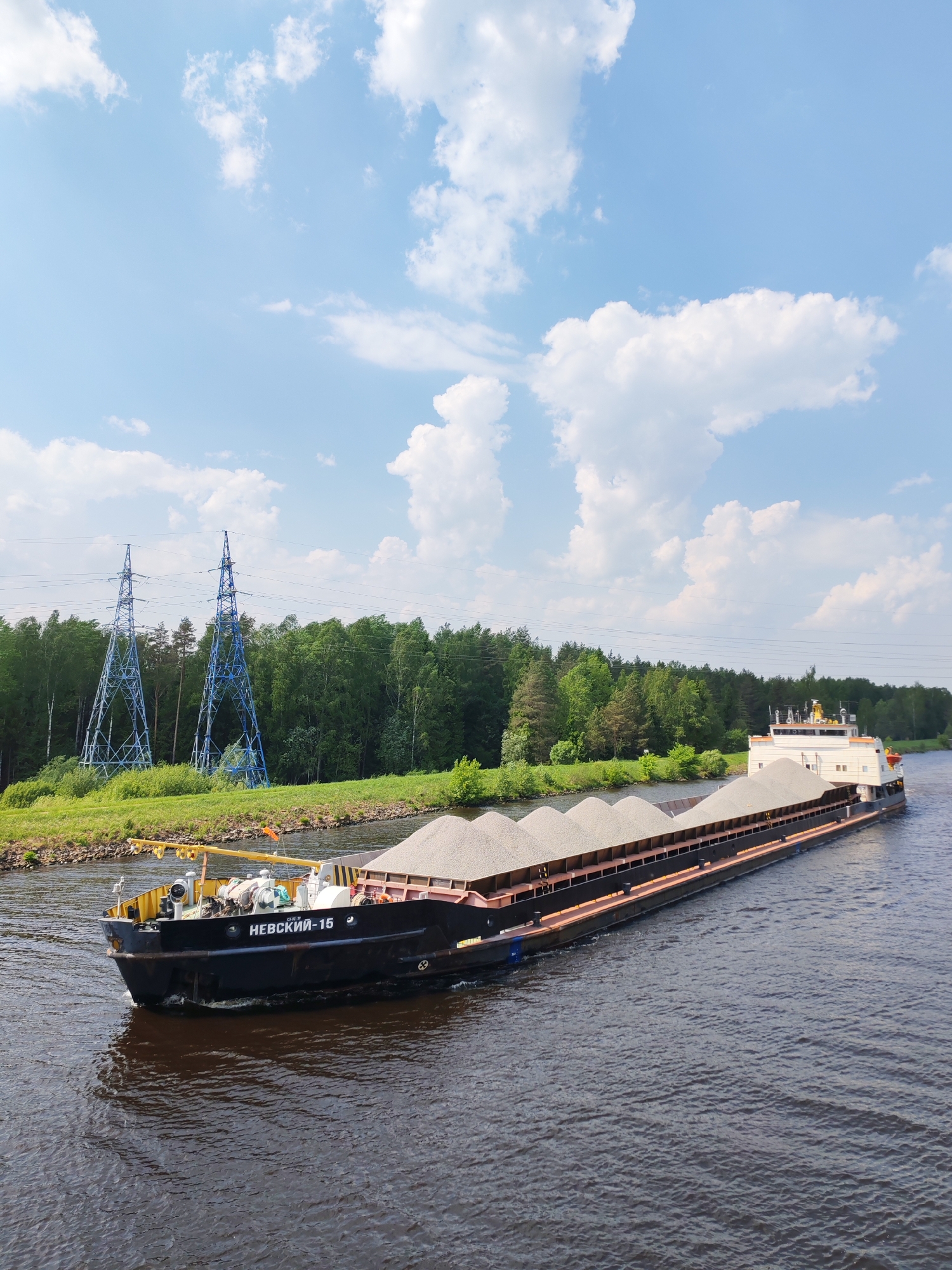 A barge, identified as "NEVSKIY 16" on its port side, is traveling along a waterway. The barge is laden with grey gravel or similar material. The surrounding environment includes green vegetation and trees along the banks, with electrical transmission towers visible in the background. The sky above is blue with scattered white clouds. The scene appears to be a waterway in a rural or suburban area, potentially near a city such as Tempy, Russia.