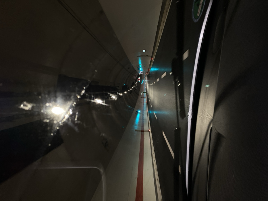 The media file captures an interior view from a moving vehicle, likely a train, as it traverses a dark, artificially lit tunnel. The perspective looks forward and slightly to the right, showcasing the tunnel's infrastructure.
On the left side of the image, the curved concrete wall of the tunnel is visible, reflecting various bright, circular white lights. These reflections appear on what seems to be a window, potentially blurred by speed or condensation, creating a shimmering effect.
Towards the center and extending into the distance, a long, narrow white pathway or platform is prominent, marked with a continuous red stripe down its middle. This pathway is illuminated by evenly spaced blue-green lights embedded along its edge, guiding the way through the darkness.
To the right, a darker, flat surface resembling a track or road runs parallel to the white pathway. This surface features intermittent white dashed lines, similar to road markings, and is also punctuated by more of the blue-green lights. The rightmost edge of the frame shows a dark, textured surface, possibly part of the train's interior or another structure within the tunnel. A green and white sticker or sign is partially visible on the upper right, but its text is illegible.
Further into the tunnel, the lights continue, diminishing in size and intensity with distance, with additional red and green signal lights observable. The environment is entirely dependent on artificial illumination, suggesting it's either nighttime or deep within an underground passage. The scene conveys a sense of forward motion through a highly structured and engineered environment. Given the location context of Pelev Brijeg, Montenegro, the tunnel is most likely a railway tunnel, part of the Belgrade–Bar railway.