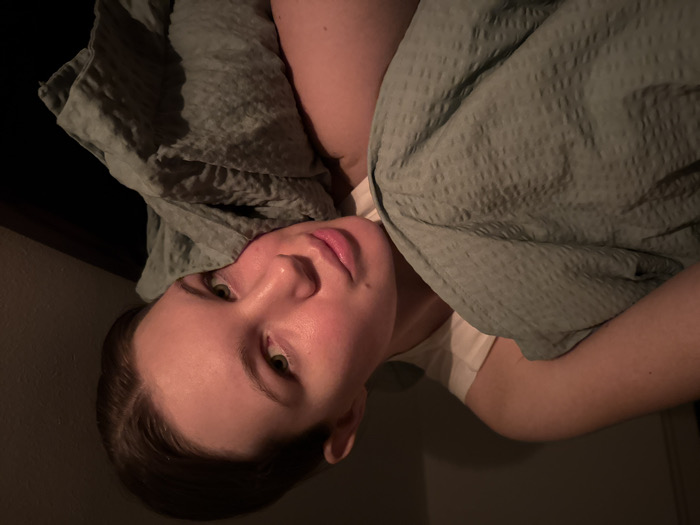 A young adult female, with light skin and dark hair, is depicted in a close-up, low-light indoor setting within the city of Vozdovac Urban Municipality, Serbia. She is positioned lying down, with her head resting on a folded section of a sage-green, textured blanket that partially covers her upper torso. A white sleeveless garment is visible beneath the blanket on her shoulder and chest. Her eyes are open, and she looks directly towards the camera. Soft, warm light illuminates her face from the left, creating subtle highlights and shadows. The background is dark and indistinct. No other individuals or prominent objects are visible.