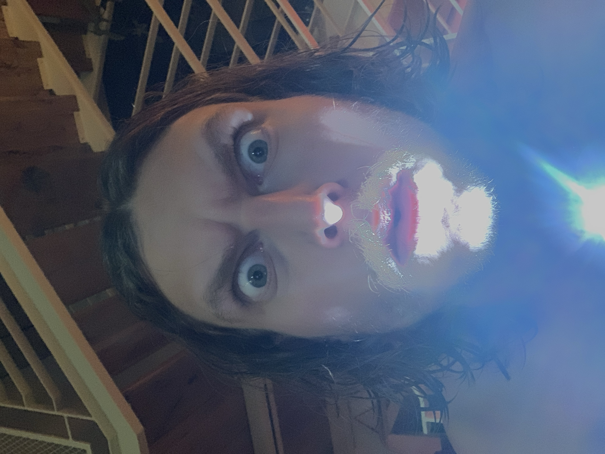 A selfie was taken at night, possibly in Tel Aviv, Israel. The person has long brown hair and light skin. The face is illuminated by a bright blue light, obscuring details and casting stark shadows. A staircase railing is partially visible in the background.