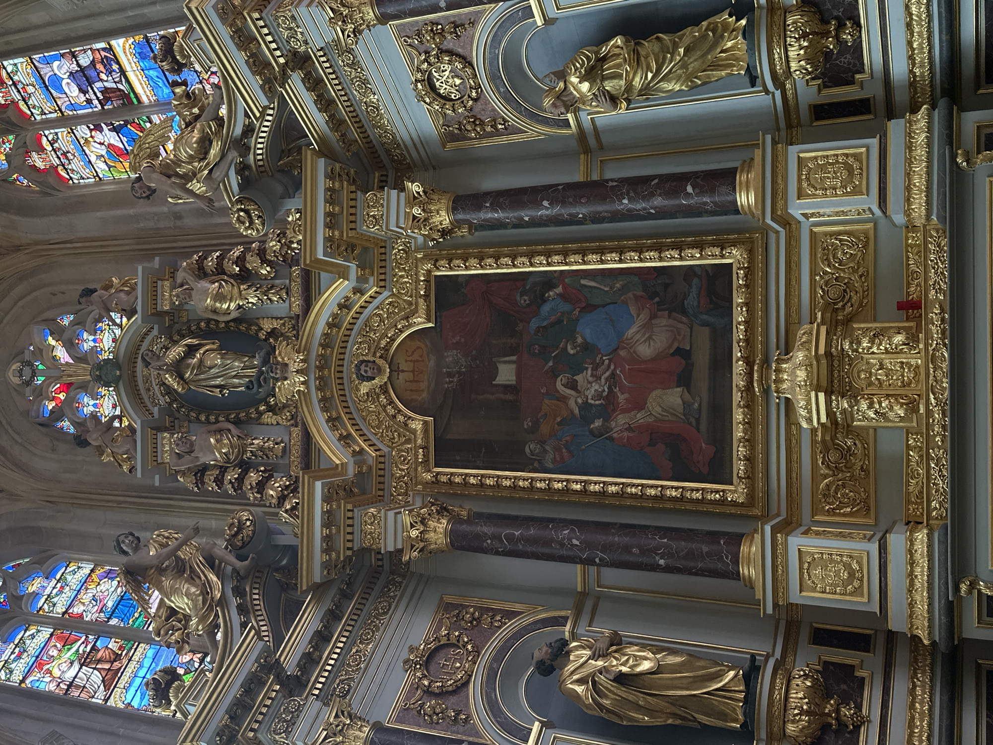 The image depicts the interior of a grand church, likely a cathedral or a prominent basilica, given the ornate architectural details and religious artwork. The focal point is a large, gilded altarpiece featuring a religious painting at its center. The painting portrays a scene with several figures, likely a biblical narrative given the religious context. Surrounding the painting are intricate gold carvings, statues, and architectural elements characteristic of Baroque or Neo-Classical styles.
On either side of the central altarpiece, at a lower level, stand large golden statues of robed figures, possibly saints or religious figures. Above these, and integrated into the upper architecture, are more gilded statues, including angelic figures with wings, and decorative elements. The background is dominated by large, colorful stained-glass windows, depicting religious scenes or motifs in vibrant hues of blue, red, and gold, typical of Gothic or Neo-Gothic architecture.
The church interior exhibits a palette of gold, deep red, marble, and muted blue-grey tones in the walls and architectural features. The lighting suggests an indoor environment, possibly with natural light filtering through the stained-glass windows and some artificial lighting. The overall impression is one of opulence, religious devotion, and historical significance. This specific altarpiece and its surrounding decorations are found in the Saint-Jacques church in Dieppe, France.