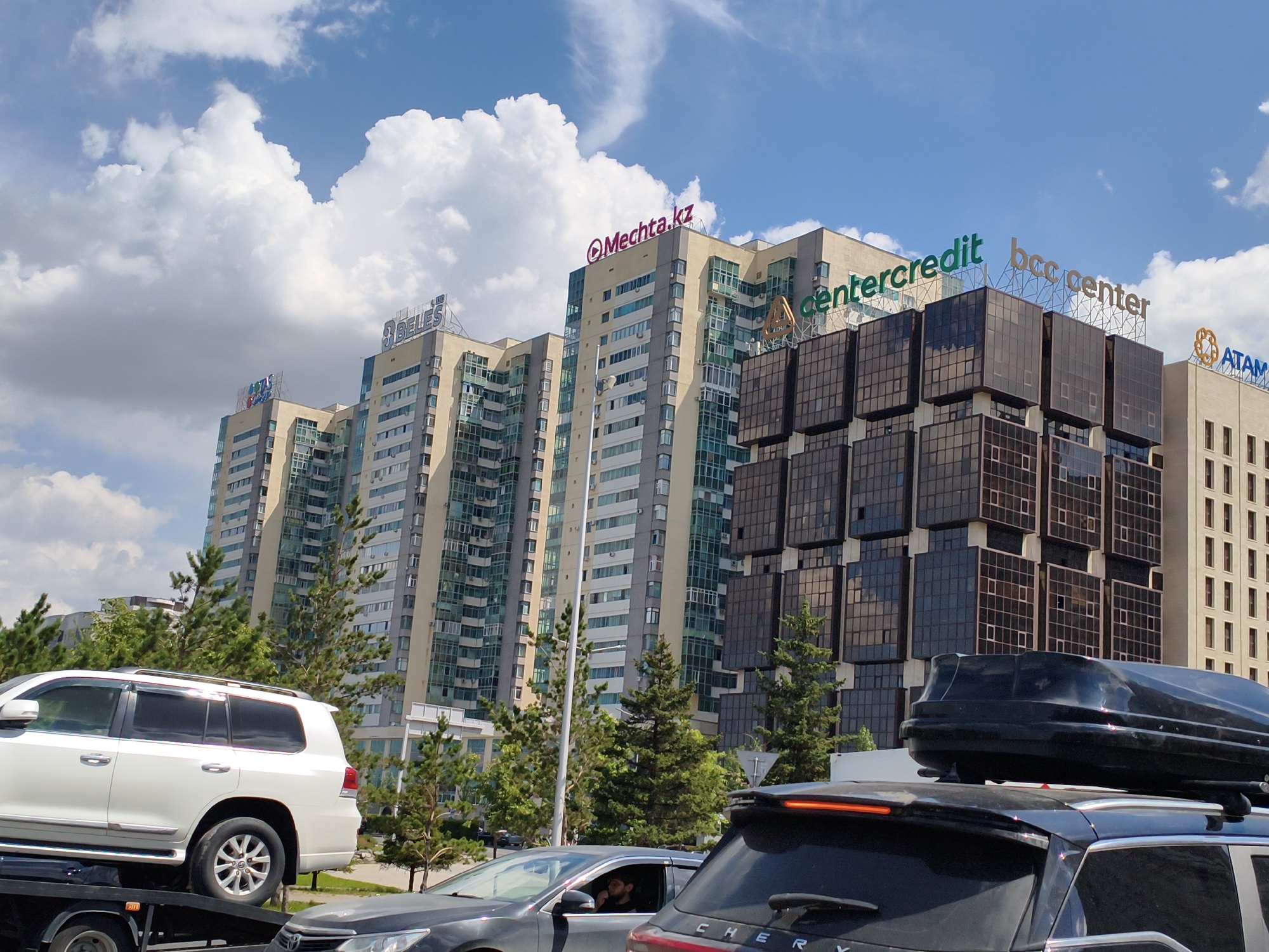 This media file captures a street scene in Astana, Kazakhstan, likely during the daytime under a partly cloudy sky. The main subjects are several vehicles, including a white SUV being transported on a flatbed truck and other cars in traffic. In the background, a series of modern high-rise buildings dominate the skyline.
Notable architectural features include tall, multi-story structures with glass facades and distinct signage. One building displays the text "Mechtа.kz" in pink, while another has "centercredit" in green and "bcc center" in orange. A third building on the far right features the text "ATAM" with an orange circular logo. These buildings suggest a commercial or business district.
The activity appears to be ordinary city traffic, with cars moving along a road. The presence of the white SUV on the flatbed truck might indicate a vehicle being moved for repair, sale, or relocation. The overall impression is one of a contemporary urban environment. The lighting suggests it is daytime, and the fluffy white clouds against the blue sky indicate fair weather.
