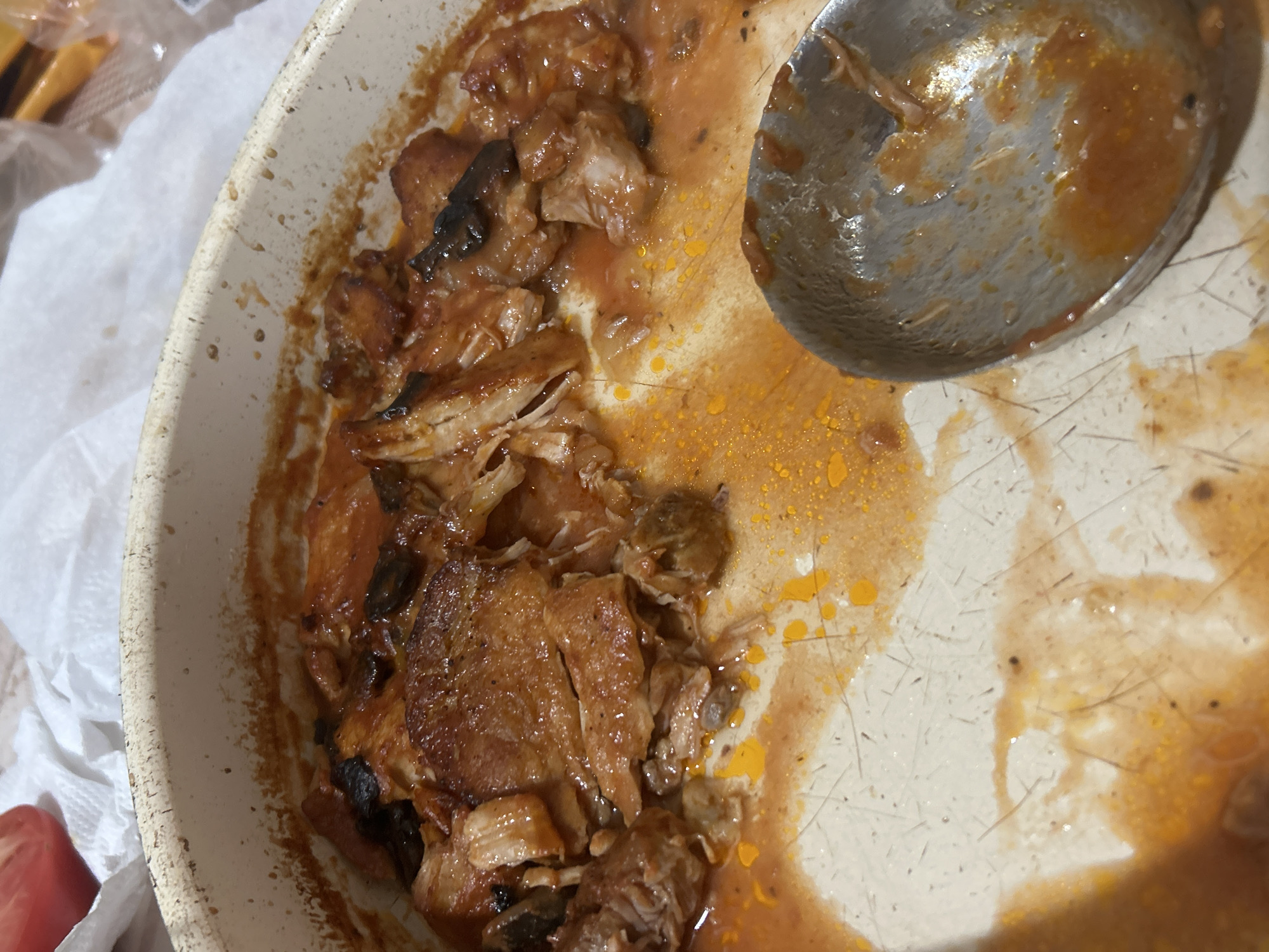 The image depicts a partially eaten meal in a ceramic pan. The food remnants appear to be some kind of meat stew with a reddish-brown sauce and possibly mushrooms. A metal spoon sits in the pan, also covered with the sauce. The pan's interior surface is scratched and stained. Around the pan are a few crumpled pieces of white paper. A section of a red object is visible to the left. The location cannot be determined from the image.