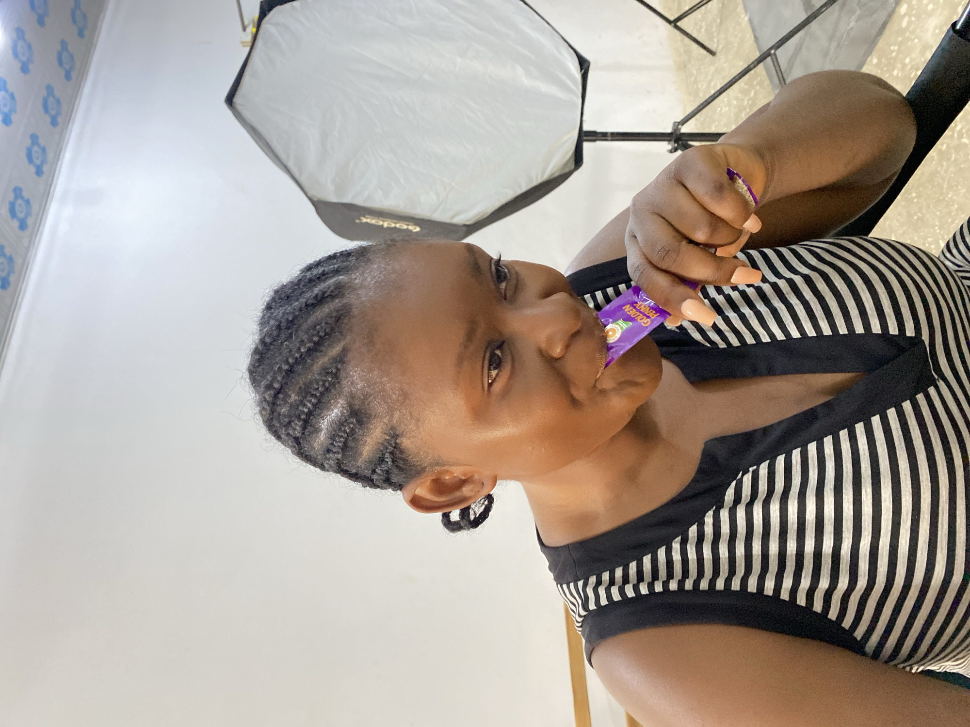 A woman with braided hair is holding a purple candy wrapper in her left hand and putting it in her mouth. She is wearing a black and white striped shirt. Behind her is a large white light diffuser and a white wall with blue patterns. The setting appears to be indoors, possibly in a studio or room with photography equipment. No specific details suggest the location is Gura Top, Nigeria.