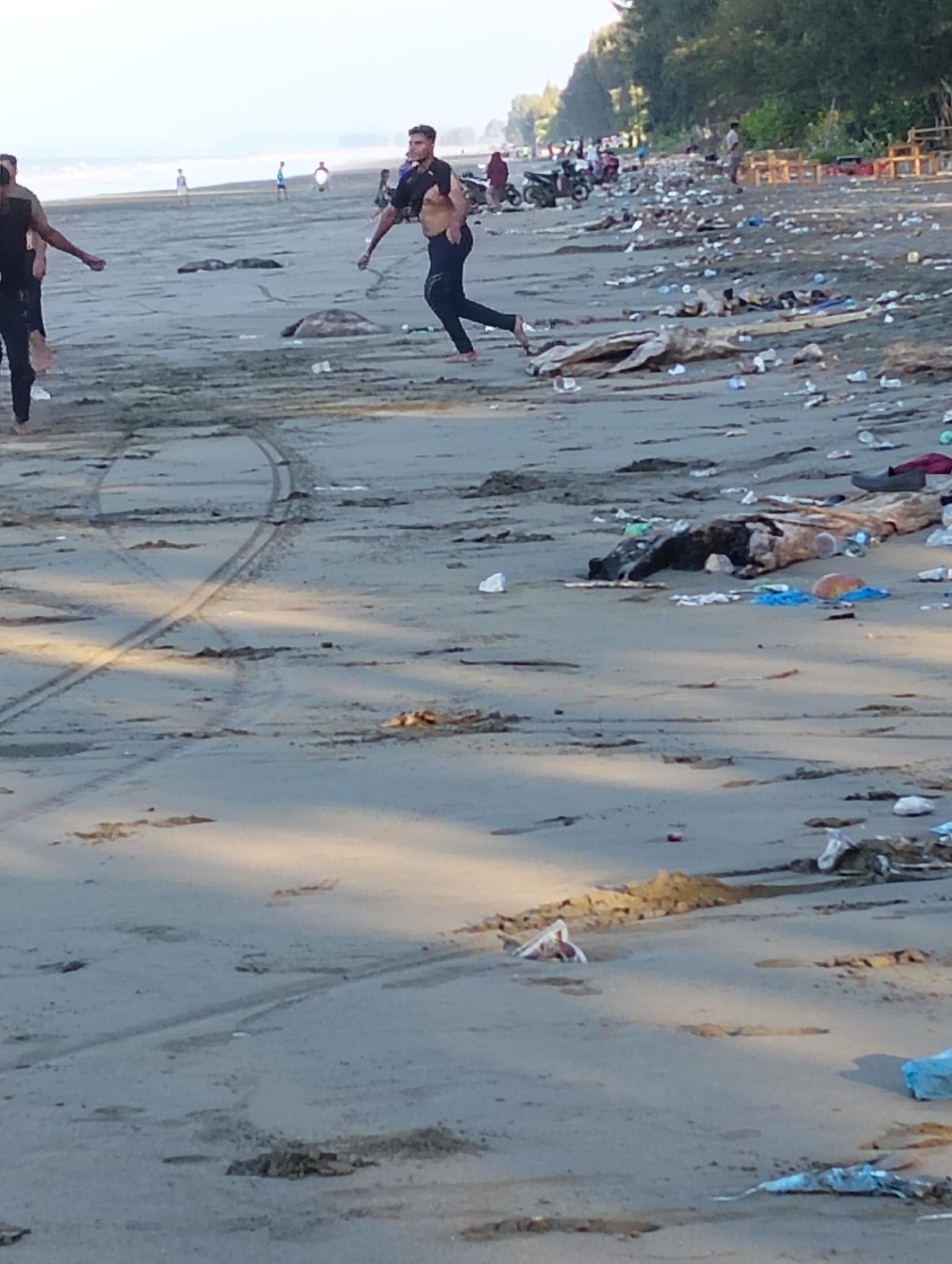 The image depicts a polluted beach in Peukan Sot, Indonesia. The sand is littered with a large amount of trash, including plastic bottles, bags, and debris. In the foreground, tire tracks are visible on the wet sand. Several people are present on the beach. In the mid-ground, two men are running, one of whom is shirtless and wearing dark pants, while the other is wearing a dark t-shirt and dark pants. In the background, there are trees along the coastline and more people, along with some motorcycles and wooden structures, suggesting a populated area. The overall scene is one of environmental neglect on a beachside.