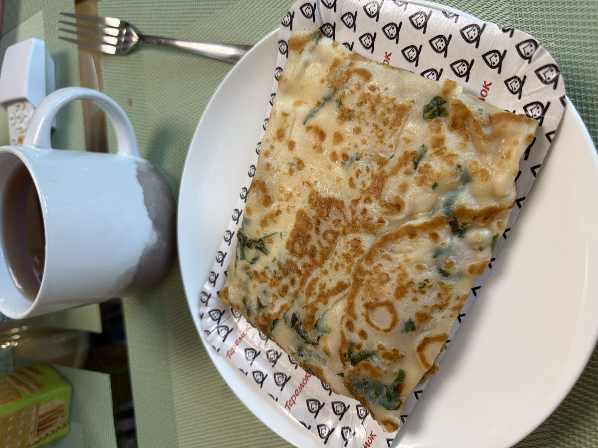 A close-up, eye-level shot captures a serving of blini with greens and cheese, presented on a white plate with a patterned paper liner that reads "Теремок" (Teremok). The blini is folded and partially covers the plate, showcasing its golden-brown, slightly crispy exterior and visible green herbs within. To the right, a silver fork rests on a green placemat. In the background, a white mug filled with dark tea sits on the same green placemat. To the left and slightly out of focus, a portion of a green and yellow box is visible. The overall setting suggests a casual meal at a cafe, possibly Teremok, a well-known chain of fast-casual restaurants in Moscow, Russia, specializing in blini. The lighting is natural, casting soft shadows and highlighting the textures of the food and tableware.