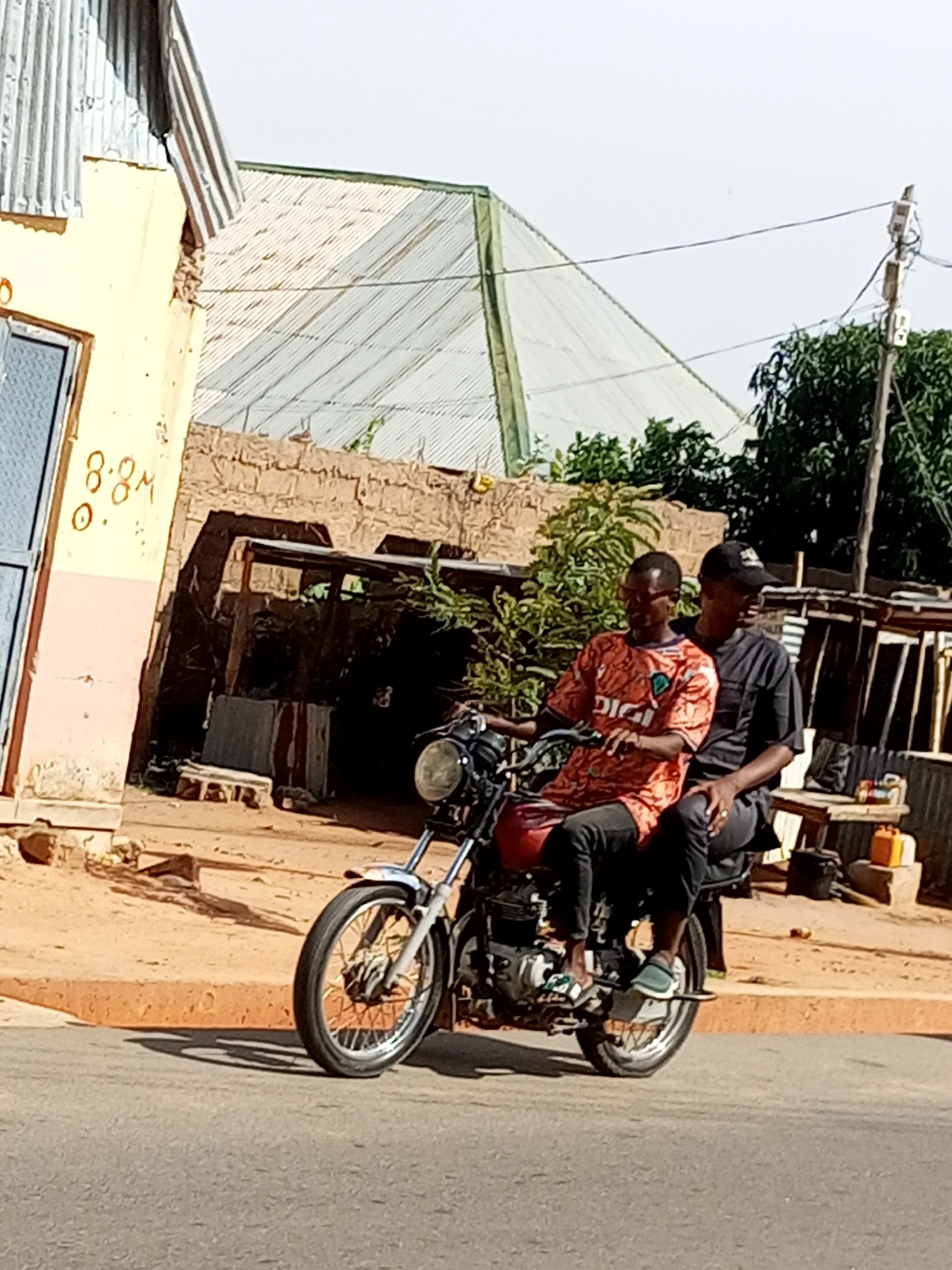 In Gombe, Nigeria, two individuals are seen riding a motorcycle. The person in the front is wearing an orange t-shirt with the word "DIGI" printed on it and dark pants. They are also wearing glasses. The person in the back is wearing a black t-shirt and a black baseball cap. They are seated behind the driver. The motorcycle is red with visible chrome parts, including the front wheel and handlebars. The background features a building with a yellow and pink facade, a corrugated metal roof, and a brick wall. Further back, there are trees and what appears to be a market stall with some goods displayed, including yellow containers and other items on a wooden surface. The ground is a mix of asphalt and dirt. The lighting suggests it is daytime, with shadows cast on the ground.