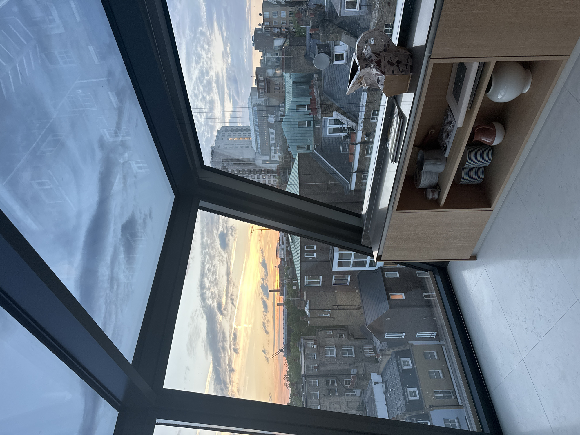 The image shows a view of the city of London, United Kingdom, through a large, modern window with a black frame. The window is composed of several triangular and rectangular panes. Outside, the cityscape is a mix of low-rise buildings, some with green or gray roofs, and taller structures in the distance. The sky is filled with clouds and hints of sunlight, suggesting either dawn or dusk.
To the right of the window, a light-colored wooden cabinet is visible. On its shelves are various objects, including a white vase, some stacked plates or bowls, a picture frame, and a decorative object wrapped in paper or fabric. The floor is tiled with light gray or white tiles. There are no people visible in the image.