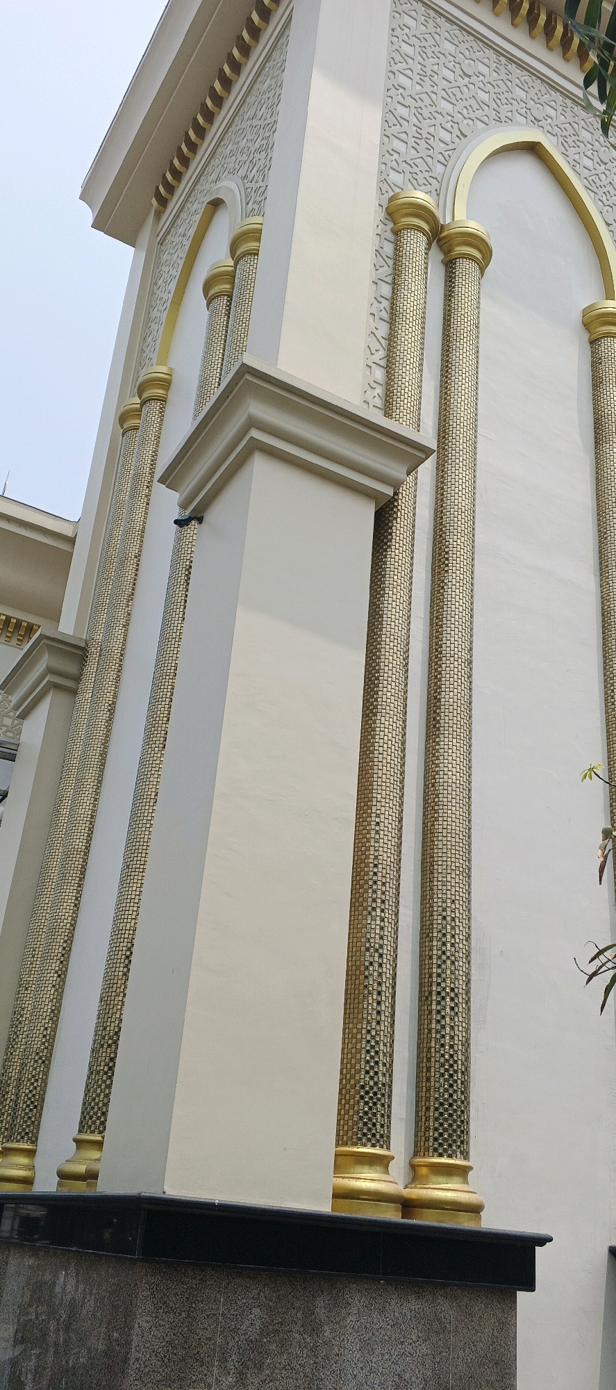 The image shows the exterior of a building, likely a mosque, with a cream-colored facade. It features several tall, cylindrical columns that are covered in gold mosaic tiles. The columns have ornate gold capitals and bases. Above the columns, there is decorative architectural detail with a geometric pattern. An arched opening is partially visible behind the columns. The lower part of the building has a dark, polished stone base. The sky is overcast. A black security camera is mounted on a cream-colored pillar. The building appears to be located in the city of Sumberwudi, Indonesia.