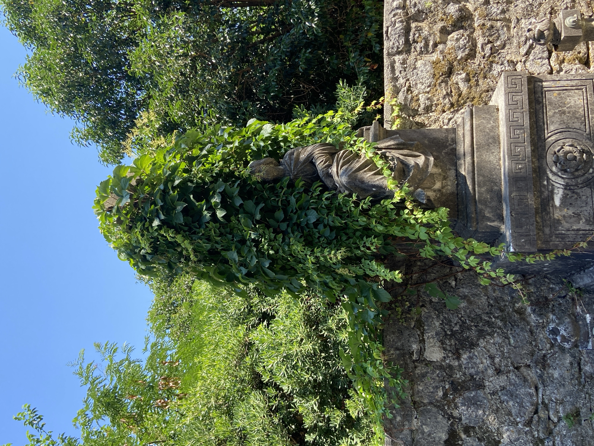 A weathered stone statue of a seated figure, mostly obscured by lush green ivy, sits atop a decorative stone pedestal. The pedestal features intricate carvings, including a Greek key pattern and a circular motif with a floral design. Behind the statue and pedestal is a rustic stone wall covered in moss. The scene is surrounded by dense green foliage and trees, set against a clear blue sky. The statue and its surroundings suggest a historical or garden setting.