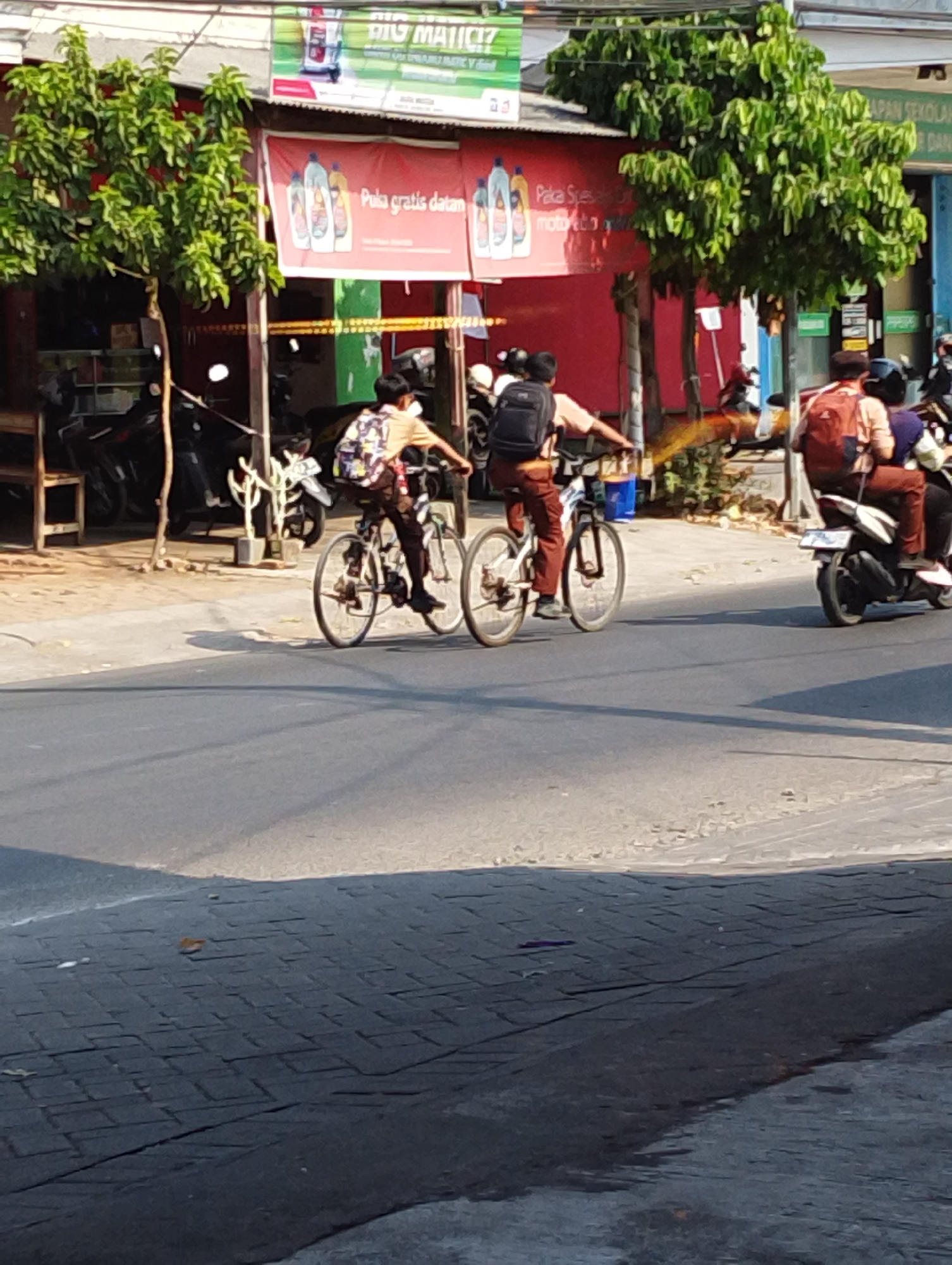 In Manaruwi, Indonesia, two school-aged children are seen riding on bicycles on a paved road. The child on the left is wearing a light brown shirt and dark pants, with a backpack on. The child on the right is wearing a similar uniform and is also carrying a backpack. Both are actively pedaling their bicycles. Behind them, a person is riding on a scooter, also with a backpack. The environment suggests a commercial area with buildings lining the street. The buildings feature advertising banners and signage. Trees with green foliage are visible along the roadside, and parked motorcycles are present near the buildings. The road itself is asphalt, and the sidewalk appears to be paved with interlocking bricks. Sunlight casts shadows on the road, indicating it is daytime.