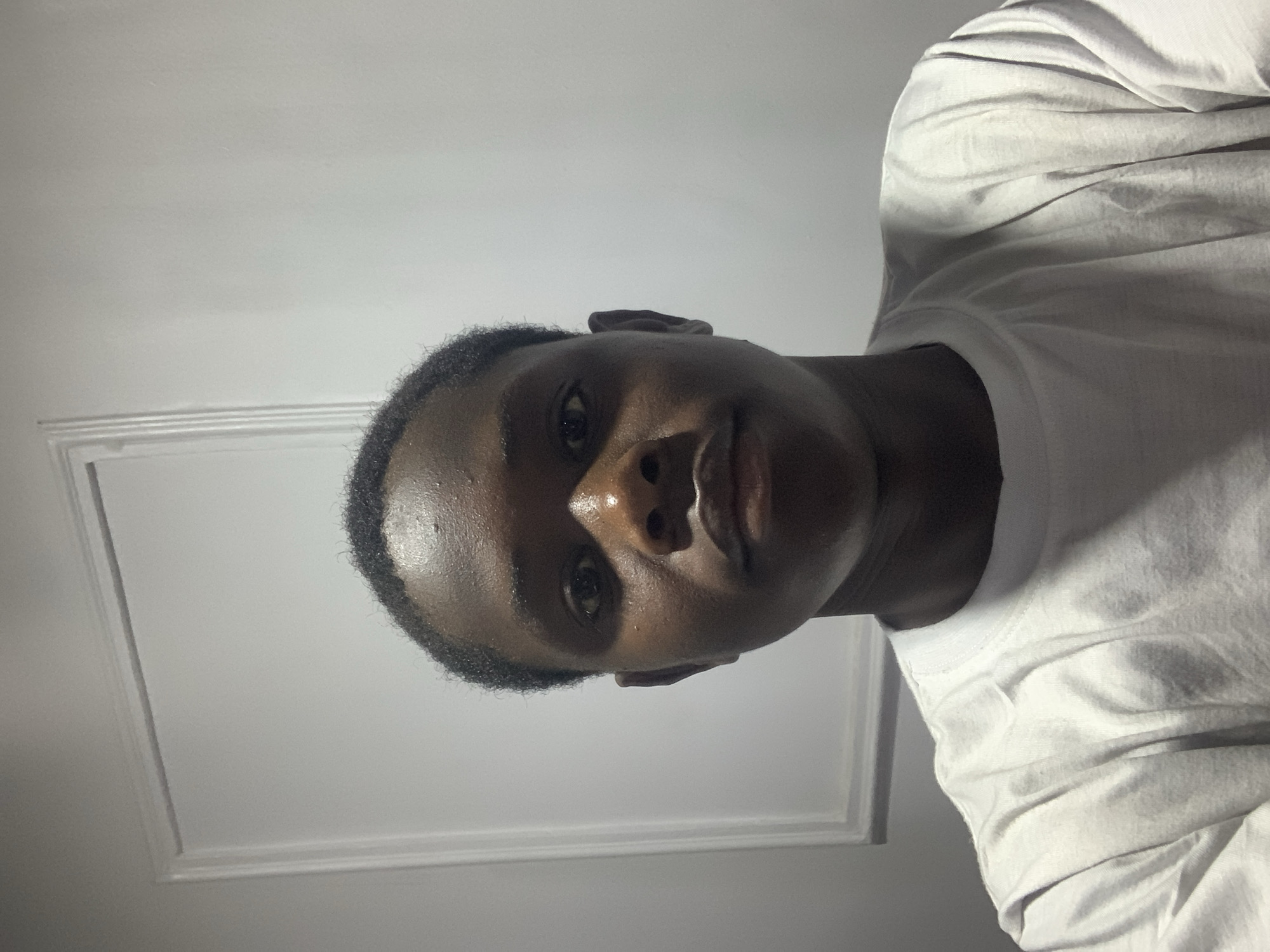 A person is seen from a low-angle perspective. The person has dark skin, short black hair, and is wearing a white t-shirt. The background is a white wall with a white square border design. There is no visible evidence to determine a specific location in Jos, Nigeria.