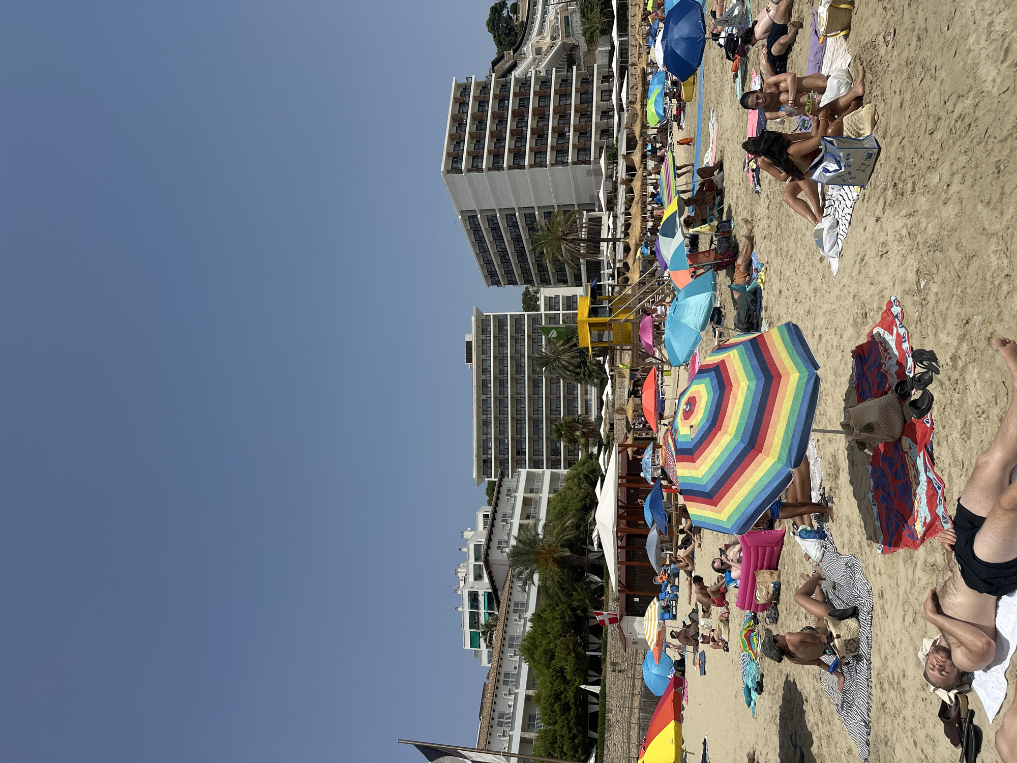 The image depicts a crowded beach scene on a sunny day, likely in the city of Sant Agustí, Spain, given the context. In the foreground, several people are relaxing on the sand, some lying down, others sitting. Beach umbrellas, in various colors including a prominent rainbow-striped one, are set up to provide shade. Beach towels and bags are scattered around. In the midground, more beachgoers are visible, along with a yellow lifeguard station and a small wooden structure with white coverings. Behind the beach, several multi-story white hotel buildings with numerous balconies line the coastline, backed by green trees and foliage. The sky is clear and blue. The overall scene conveys a lively and typical summer beach atmosphere.