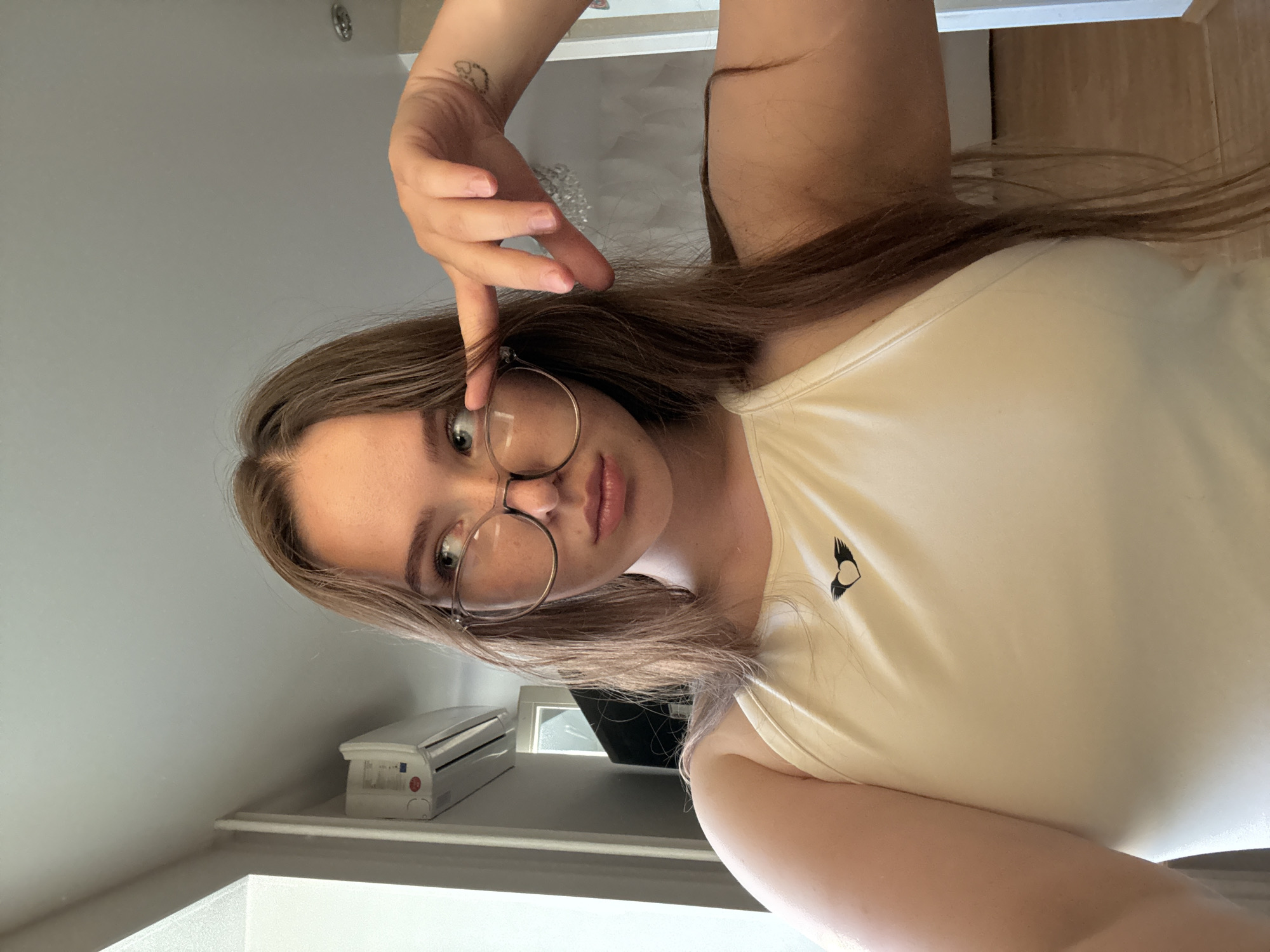 The image shows a young woman with long, light brown hair and glasses. She is wearing a light-colored, sleeveless top with a small black logo of a heart with wings on the left side of her chest. Her right arm is raised, and her hand is near her face, with a tattoo visible on her forearm. She is looking at the camera with a neutral expression. The background appears to be an indoor setting with light gray walls and a white shelf. A small white object resembling an air conditioner unit is visible on the shelf. The lighting suggests it is daytime.