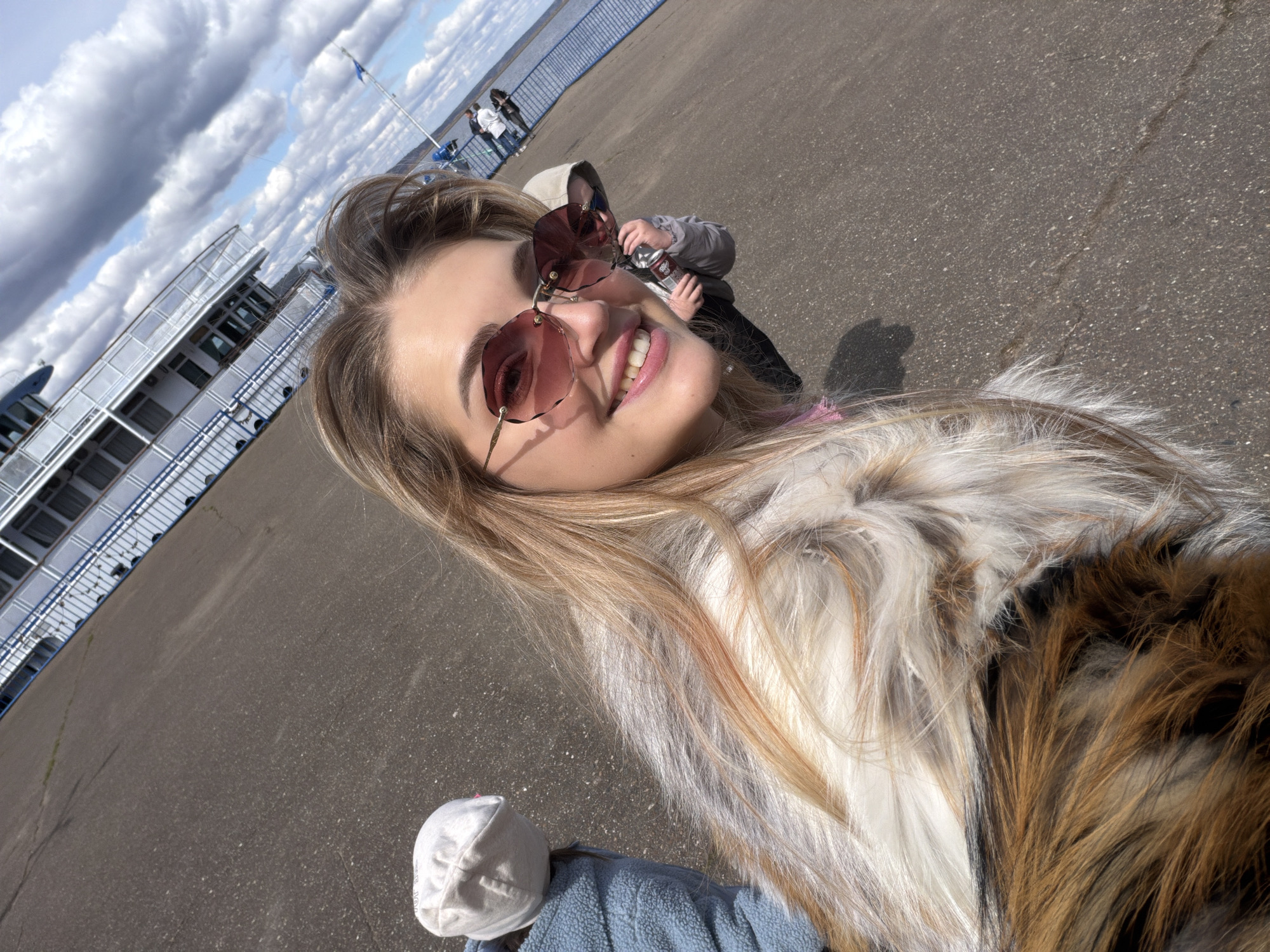 A selfie captures a smiling woman wearing sunglasses and a furry coat. Behind her, a child in sunglasses holds a can. In the background, blurred figures stand near a blue metal railing and a body of water. To the left, a section of a multi-decked ship is visible under a sky with fluffy clouds. A white hat and blue jacket lie in the foreground. The location is Chaikovsky, Russia, given the query context.
