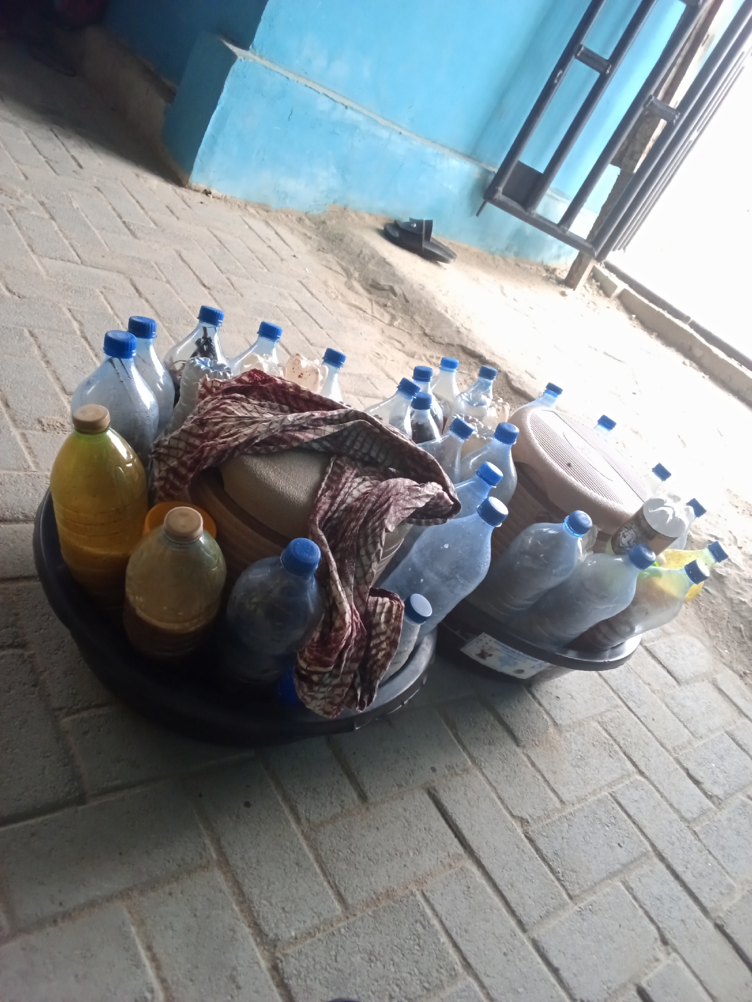 In Ado, Nigeria, a close-up, overhead view captures a collection of plastic bottles filled with various liquids, predominantly water and some yellow or light brown substances, resting in a large black basin. The bottles are mostly clear with blue caps, though some appear opaque and a few have wooden or light-colored caps. A patterned fabric, predominantly red and white, is draped over some of the bottles and a light brown, woven circular object in the center. The basin is positioned on a light gray brick or paver ground. In the background, a blue painted wall is visible, with a section of metal shelving. The overall lighting suggests an outdoor setting.