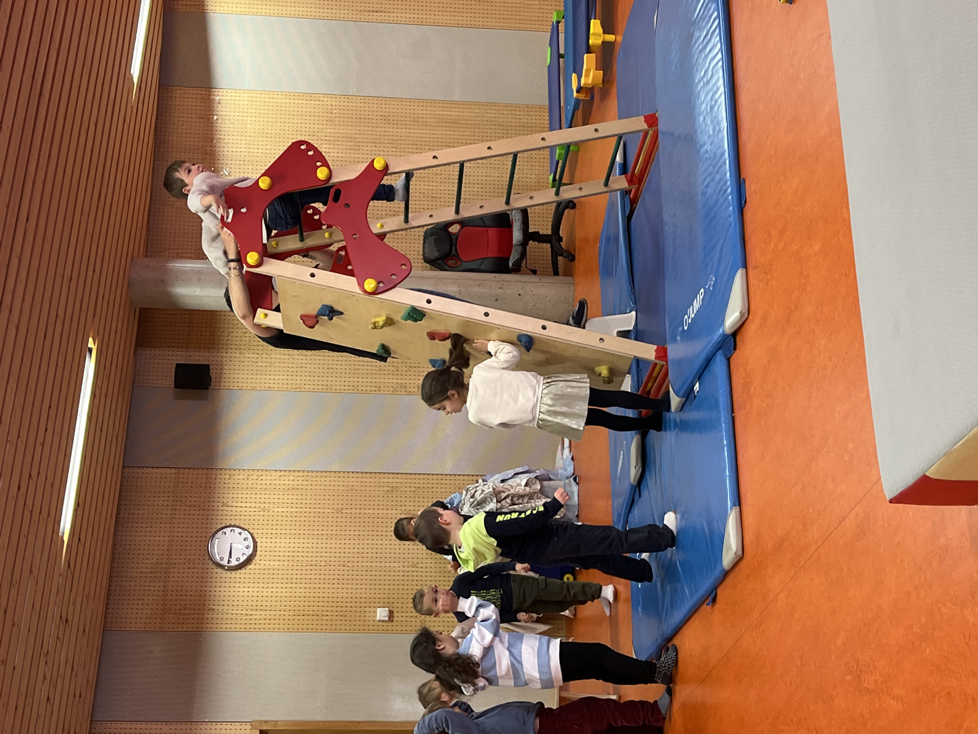 A group of children are engaged in play in an indoor gymnasium. The gymnasium has orange flooring and walls with wood paneling and a light gray section. A clock on the wall shows the time to be around 9:50.
A climbing structure is positioned on a blue gymnastics mat. One child is climbing the structure, assisted by an adult. Several other children are standing or lying on the mat, appearing to be waiting their turn or observing the activity. There's a ladder and other gymnastic equipment near the climbing structure.
The location is presumed to be in La Fouillouse, France.
