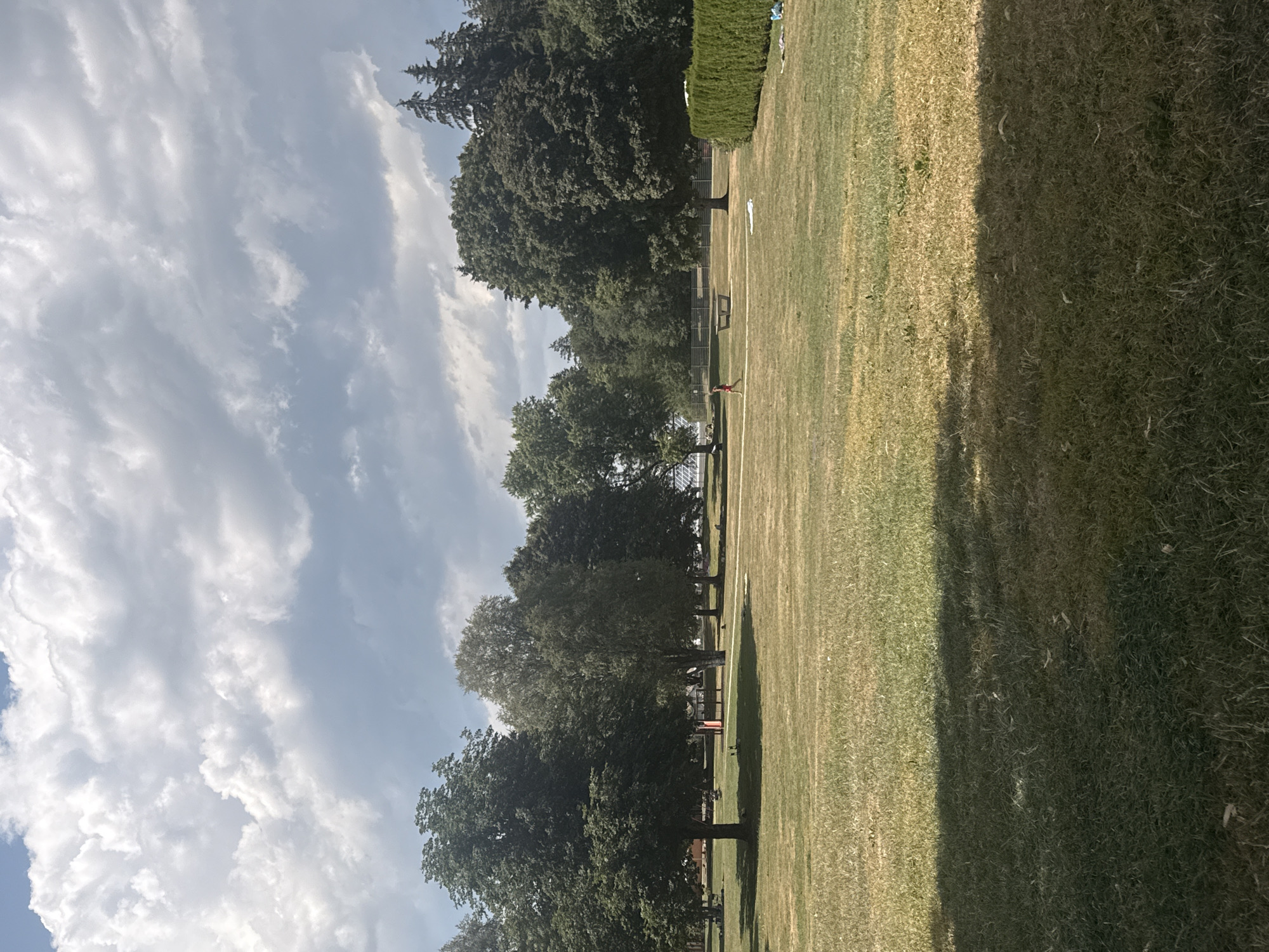The image depicts a wide, grassy park area under a dramatic, cloudy sky. The park is bordered by a dense line of green bushes on the right and a variety of mature trees on the left and in the background. In the middle ground, there are several people scattered across the lawn. One person is walking in the middle of the park, wearing a red shirt and dark shorts. Another person appears to be lying on a white blanket in the grass to the right of the walker. In the background, a bench is visible, and further back, structures and more trees suggest the edge of a developed area. The grass appears dry in some patches, indicating a sunny or dry period. This scene is located in the Parc des Crêtes in Le Locle, Switzerland, a public space known for its expansive lawns and recreational areas.