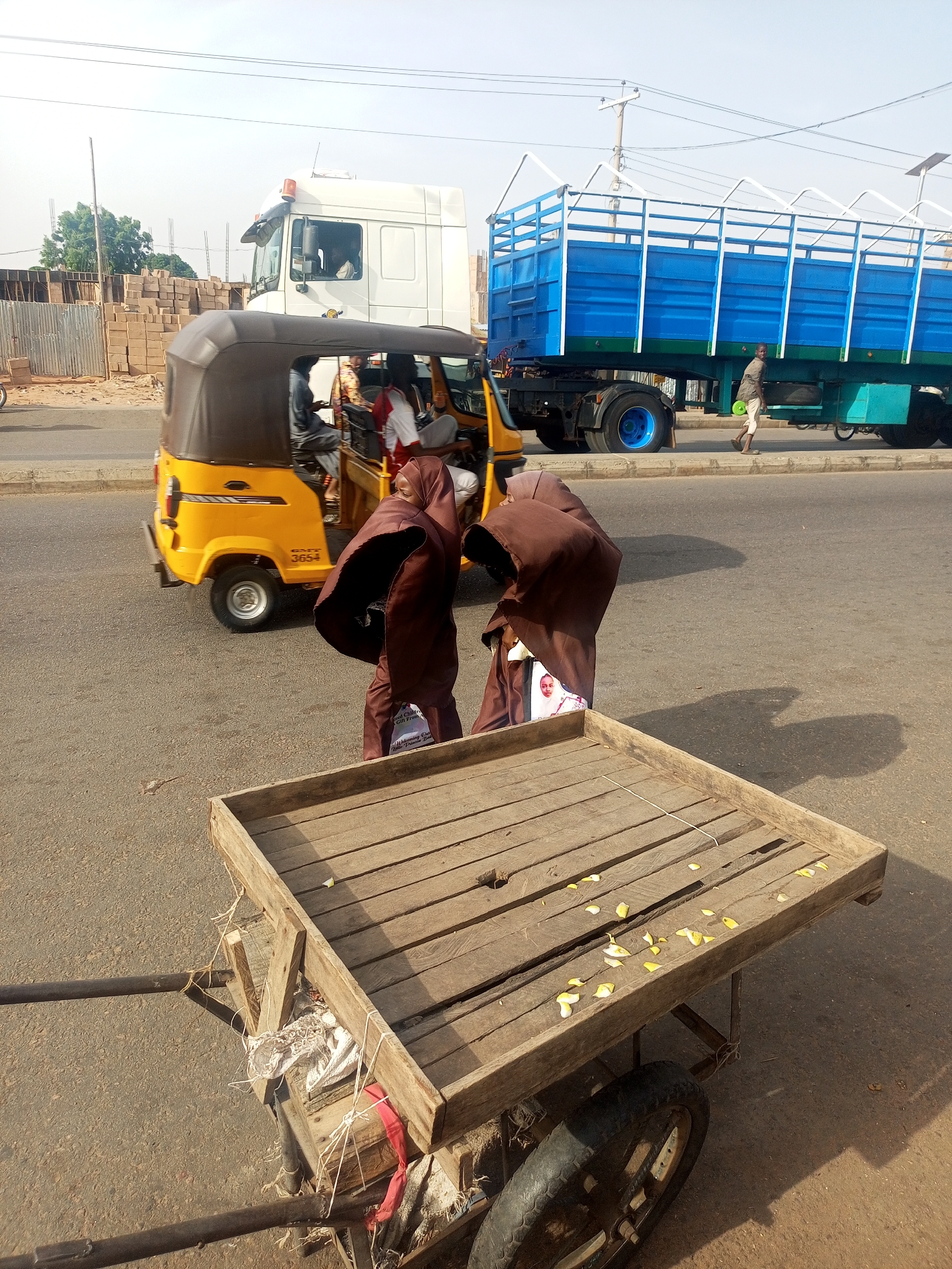 The image shows a street scene in what appears to be a city in Nigeria. In the foreground is a wooden cart with two wheels, scattered with yellow flower petals. Behind the cart, two individuals wearing brown hooded garments stand, their faces obscured. To the left, a yellow three-wheeled vehicle (possibly an auto-rickshaw or "keke") is partially visible, with passengers inside. Further back, a white truck with a visible driver is present. To the right, there's a blue flatbed truck, with a person walking on the sidewalk beside it. In the background, there are buildings and what appears to be construction materials. The road is paved and shows signs of use. Based on the context provided, the location is Tempure, Nigeria.