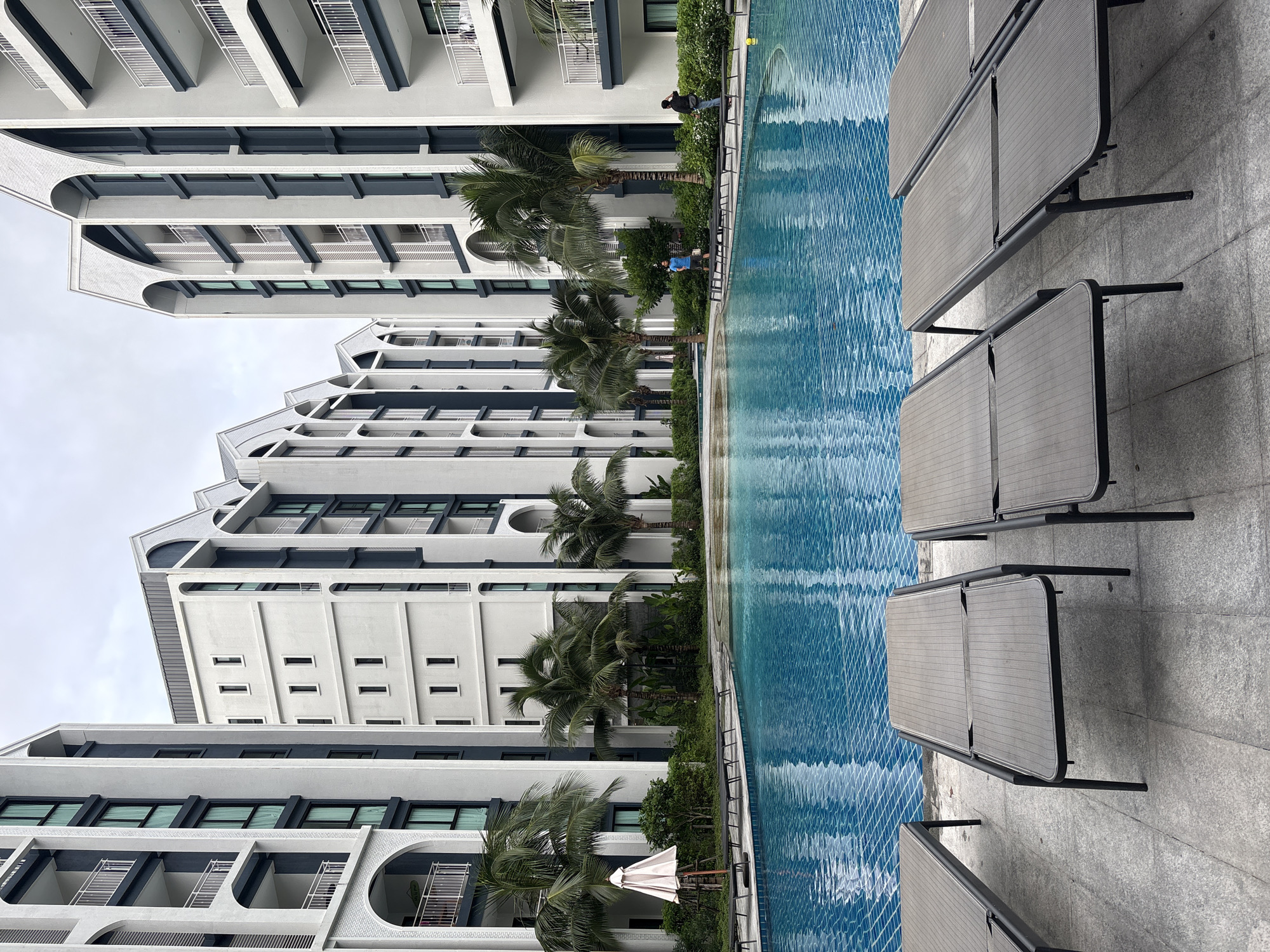 A wide-angle, eye-level shot captures a resort swimming pool area in Sakhu, Thailand. The pool, filled with bright blue water and a visible tiled bottom, occupies the center of the frame. The surrounding paved area features rows of empty, dark gray lounge chairs. Flanking the pool are modern, multi-story apartment buildings with white facades, dark gray accents, and arched window details. Lush green palm trees and other tropical foliage are visible between the buildings and around the pool area. In the background, two men are standing near the edge of the pool, and a white umbrella is positioned near the lounge chairs on the left side of the image. The sky is overcast and appears to be a light gray color.