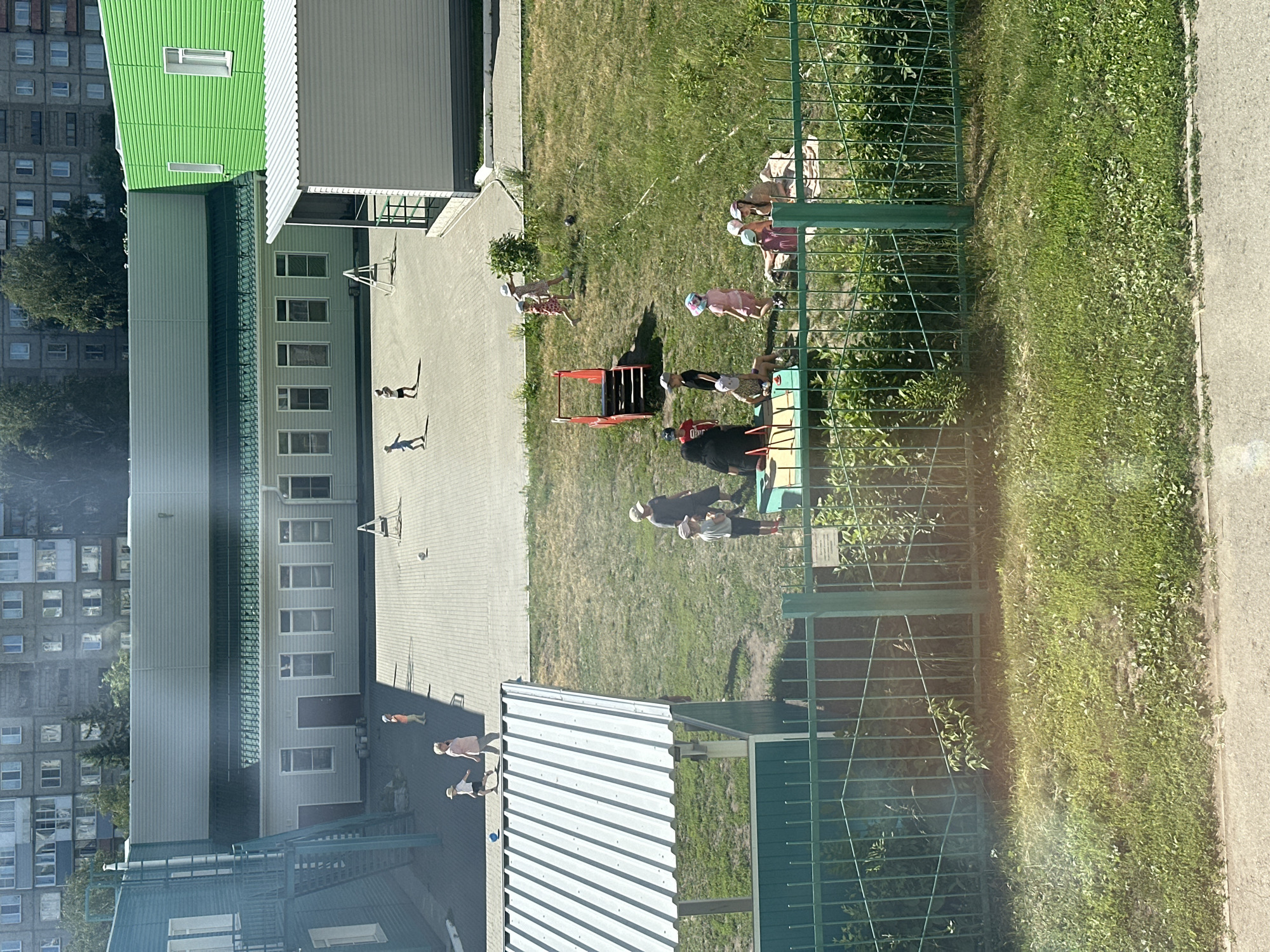 This is an aerial, medium-distance, wide shot of a kindergarten or schoolyard in Novoaltaysk, Russia, on a sunny day. A group of young children and some adults are scattered across a paved area and a grassy section, some engaging in play or conversation. The scene is framed by a green metal fence in the foreground and residential buildings in the background.
In the paved area, several children are visible. Two children are walking towards the left, one wearing an orange shirt and the other a white shirt. Another child is running in the upper left quadrant. A light-colored ball is on the ground in the middle of the paved area. Further to the right, two children are standing, one with a white hat.
The grassy area features a red and white playground structure. A group of children and at least one adult are gathered around a mat on the grass to the right of the playground. Some children are sitting, while others are standing. One child in a pink dress and hat is walking away from the playground. A dark-haired adult wearing a black top appears to be interacting with some of the children. Another adult, wearing a pink shirt, is walking on the paved area.
The environment includes a large, light-colored building with a green roof and several windows, and a bright green building. A multi-story residential building with many windows is visible in the background. The foreground is dominated by a green metal fence with diamond-shaped accents, and a patch of green grass and some foliage. The overall lighting suggests bright sunlight, with distinct shadows cast on the paved area.