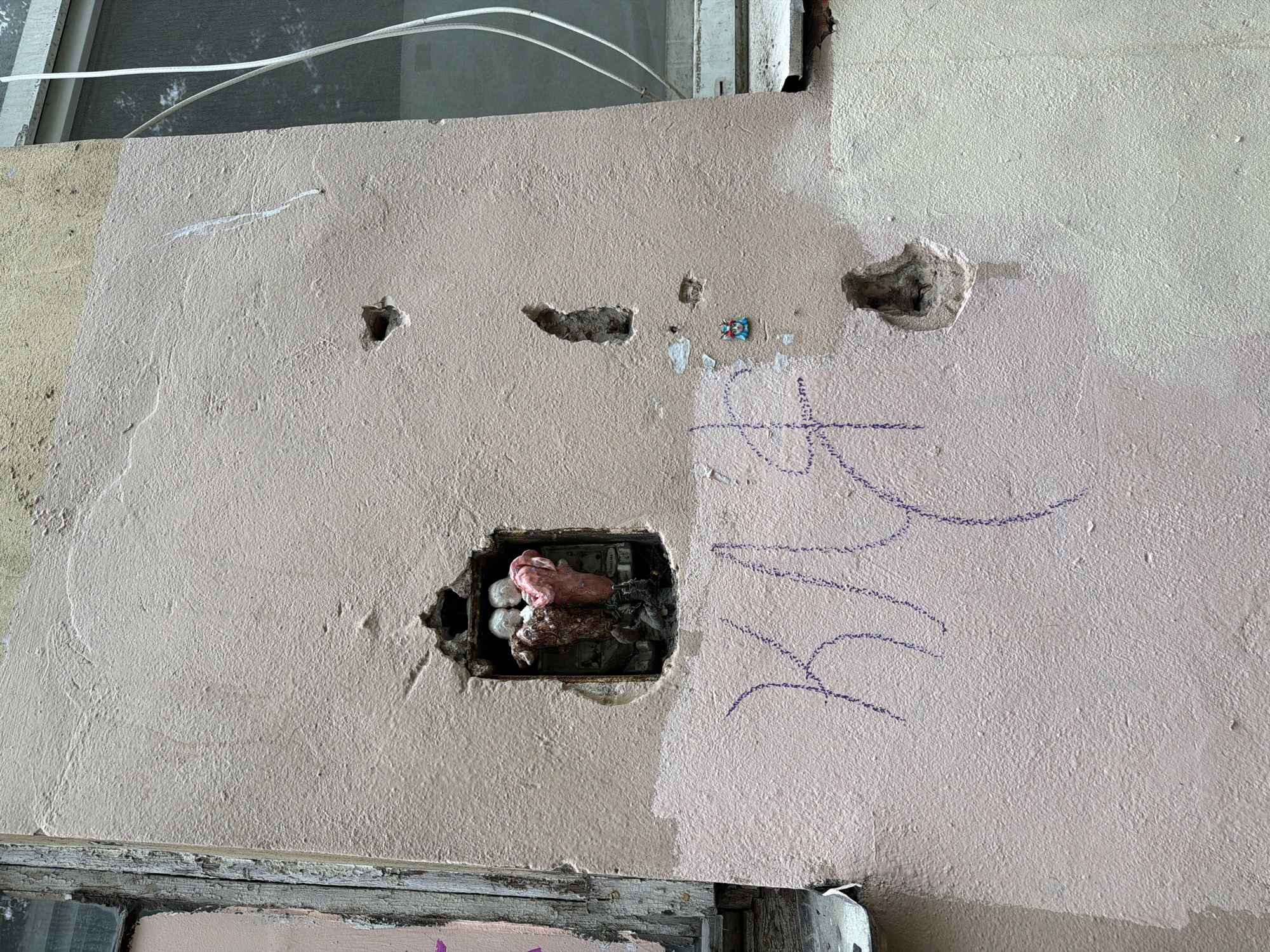 A close-up shot reveals a wall with several holes and markings. A large, rectangular hole exposes electrical wiring encased in pink and white putty or insulation. Three smaller holes are visible above this larger opening. There is blue graffiti of an unrecognizable figure to the right of the larger hole. A small, multi-colored, possibly ceramic square is located near the upper central portion of the wall. Two colors of paint are applied to the wall, separated by a vertical line. The top of a window and white electrical cables are located in the upper left portion of the shot, and a wooden window frame is at the bottom. Based on the style of the building, the scene may be located in Saint Petersburg, Russia.