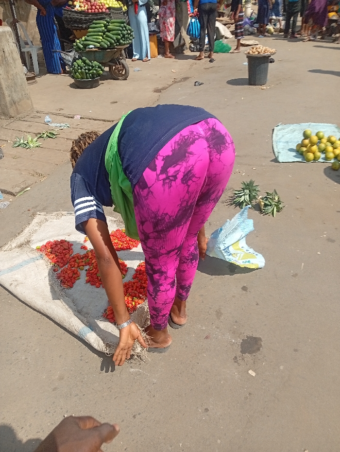 A woman in pink and black tie-dye leggings bends over a white cloth covered with red peppers on a street. She wears a navy blue shirt with white stripes on the sleeves and a light green cloth around her neck. Her hands are outstretched, one near the peppers, the other near some dried roots on the ground. Behind her, there are fruits and vegetables for sale in the open market. Some people are seen standing around. A bag and some pineapples lie on the ground nearby. A partial hand is visible in the bottom left of the frame.
