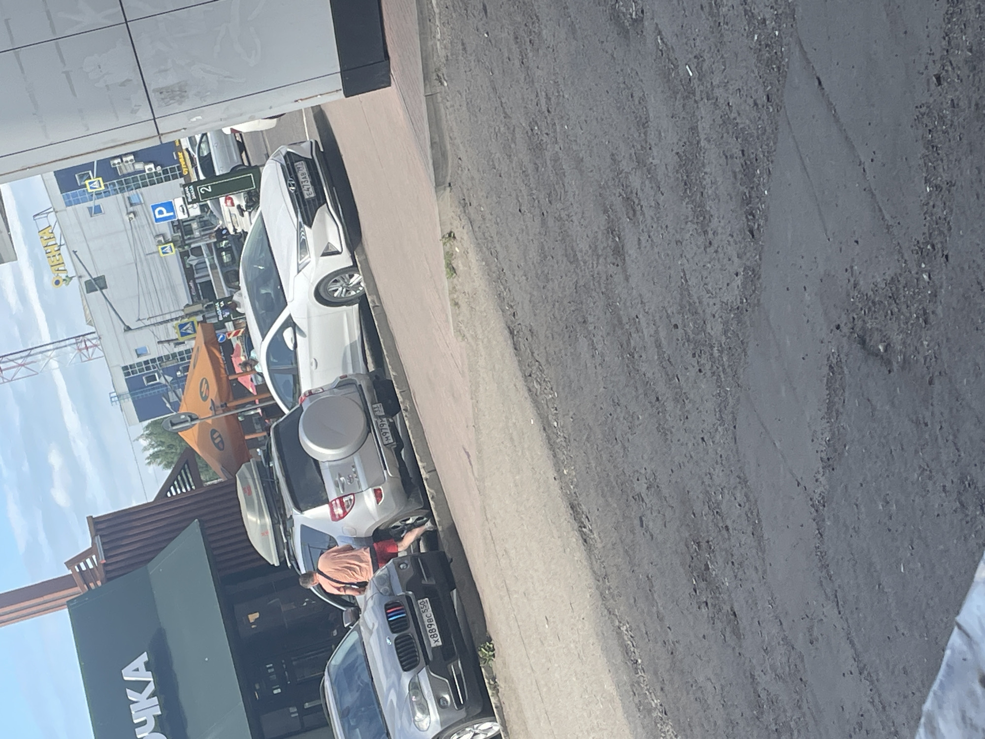 The image depicts a street scene in Zhukovo, Russia. In the foreground, a gray asphalt road with visible wear and tear is present. To the right, a building with a white and gray facade is partially visible. Adjacent to it, a sidewalk made of light-colored paving stones lines the street.
Several cars are parked on the side of the road. A silver BMW X5, with license plate "X889EC 550," is parked on the left. To its right is a silver Toyota RAV4 with a roof box and a spare tire mounted on the back, bearing license plate "M979HH 31." A man in an orange t-shirt and red shorts is seen partially obscured by the RAV4, appearing to interact with the driver's side of the vehicle. Further to the right, a white Hyundai Elantra, with license plate "E473AB 797," is parked. Beyond these cars, more vehicles are visible, including a white car and a dark SUV.
In the background, a green building with a sign featuring white text and the logo of "PYCKA" is visible. An orange awning is set up in front of a brown wooden structure. A tall metal tower is also prominent in the background. Several blue buildings with windows and signs, including one with a yellow "ЛЕНТА" sign, are situated further in the distance. Traffic signs, including a blue "P" for parking and a sign with the number "2," are also present. The sky is clear with some clouds.