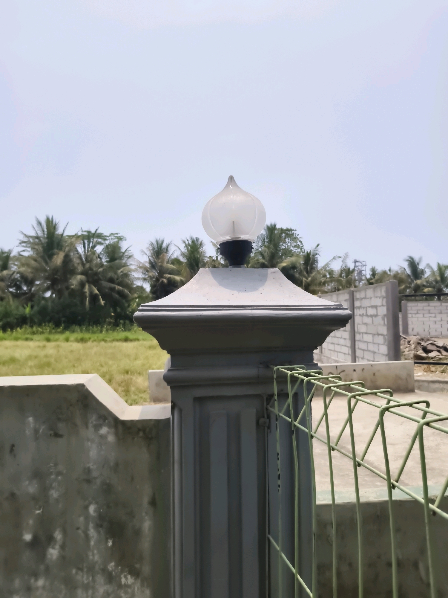 A close-up, eye-level shot showcases a grey pillar with a decorative light fixture on top, positioned in front of a rural landscape. The pillar, with its fluted design and tiered cap, stands on a concrete base. To the right, a light green wire mesh fence extends into the frame. In the background, a field of dry grass meets a line of palm trees, under a clear, bright sky. Further back, a partially constructed building with concrete block walls is visible, suggesting ongoing construction in the area. The scene appears to be an outdoor setting in Pulosampel, Indonesia, with evidence of both natural and man-made elements.