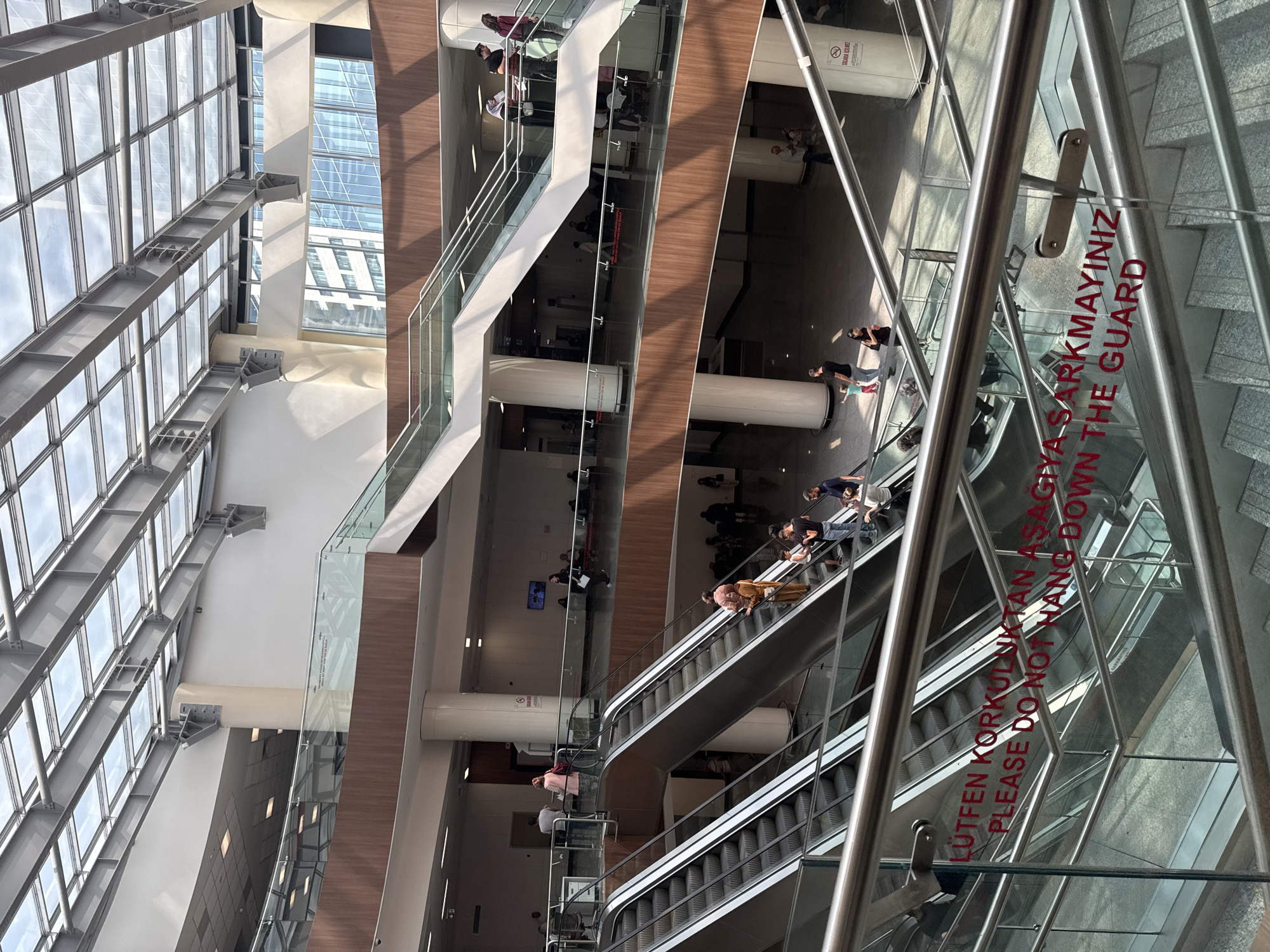 An interior shot captures a modern, multi-level atrium with a glass ceiling and steel beams. Several escalators and staircases, framed by glass and metal railings, connect the different floors. People are seen using the escalators, walking, and standing in various areas. Red text on a glass railing reads "LÜTFEN KORKULUKTAN AŞAĞIYA SARKMAYINIZ PLEASE DO NOT HANG DOWN THE GUARD." The scene suggests a public building, possibly a hospital or a large office complex, located in Çankaya, Turkey, given the Turkish text.