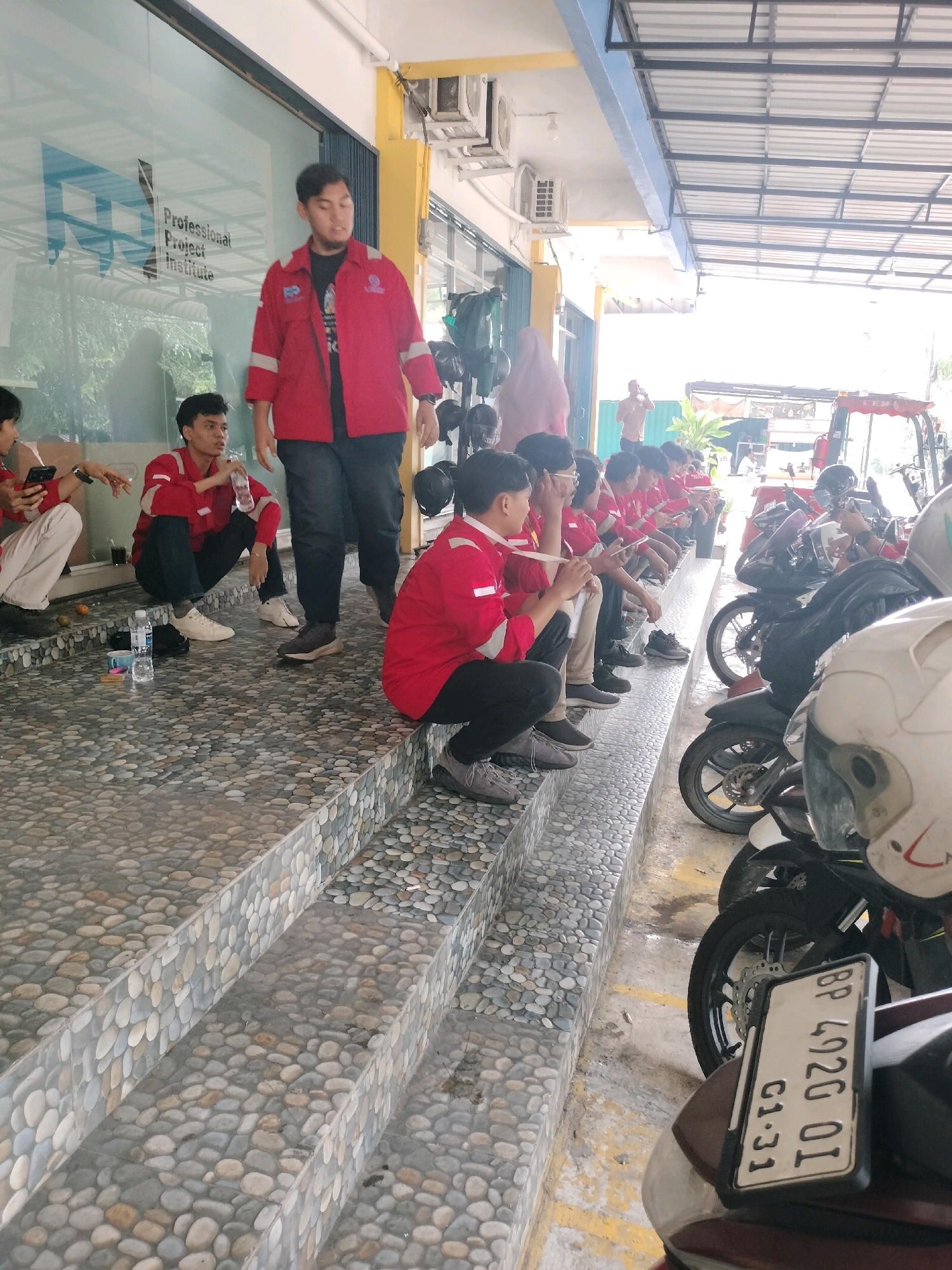 In Batam, Indonesia, a group of young men in red jackets, likely students or participants in a program at the Professional Project Institute, are gathered on a set of pebble-tiled steps. Some are seated, others stand, and one individual is walking away from the camera. A few are looking at their phones or holding water bottles. In the background, motorcycles are parked, and a red forklift is visible. The scene appears to be an outdoor area adjacent to a building with large windows and a covered walkway. The atmosphere suggests a casual break or waiting period.