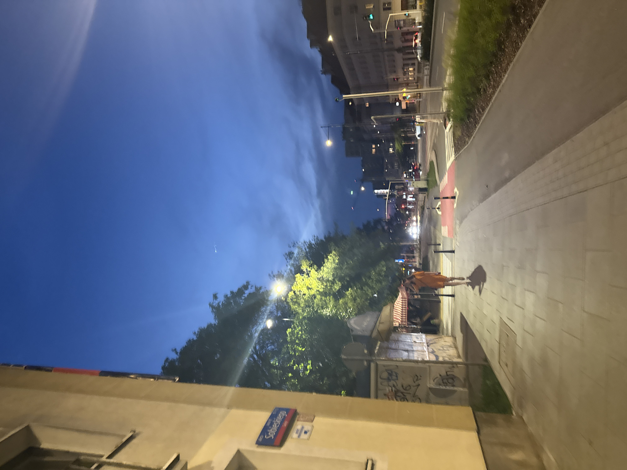 The image depicts a nighttime scene on a city street in Unknown, Unknown. A solitary person, wearing an orange outfit, walks along a sidewalk. To the left, a building with yellow siding features a street sign reading "Sobieskiego". Further down the street, there are several businesses with striped awnings, and graffiti adorns some of the structures. The street itself has traffic lights and several cars. The sky above is a deep blue, with clouds visible and some streetlights illuminating the scene.