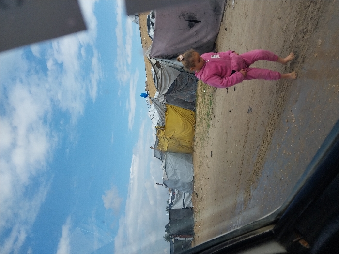 The image depicts an informal settlement in Khan Yunis, Palestinian Territory, under a partly cloudy blue sky. A young girl, appearing to be between 5-8 years old, is walking barefoot on a muddy dirt road that shows tire tracks. She wears a solid pink tracksuit with a hooded top and matching pants, and her light-colored hair is pulled back. Her gaze is directed downward. To her left and behind her, a series of makeshift shelters or tents are visible. These structures are constructed from various materials including grey, yellow, and dark-colored tarpaulins or fabrics, some appearing to be draped over frames or damaged. The photograph is taken from inside a vehicle, with a portion of the car's window frame and interior visible in the lower left and a blurred reflection in the upper right.