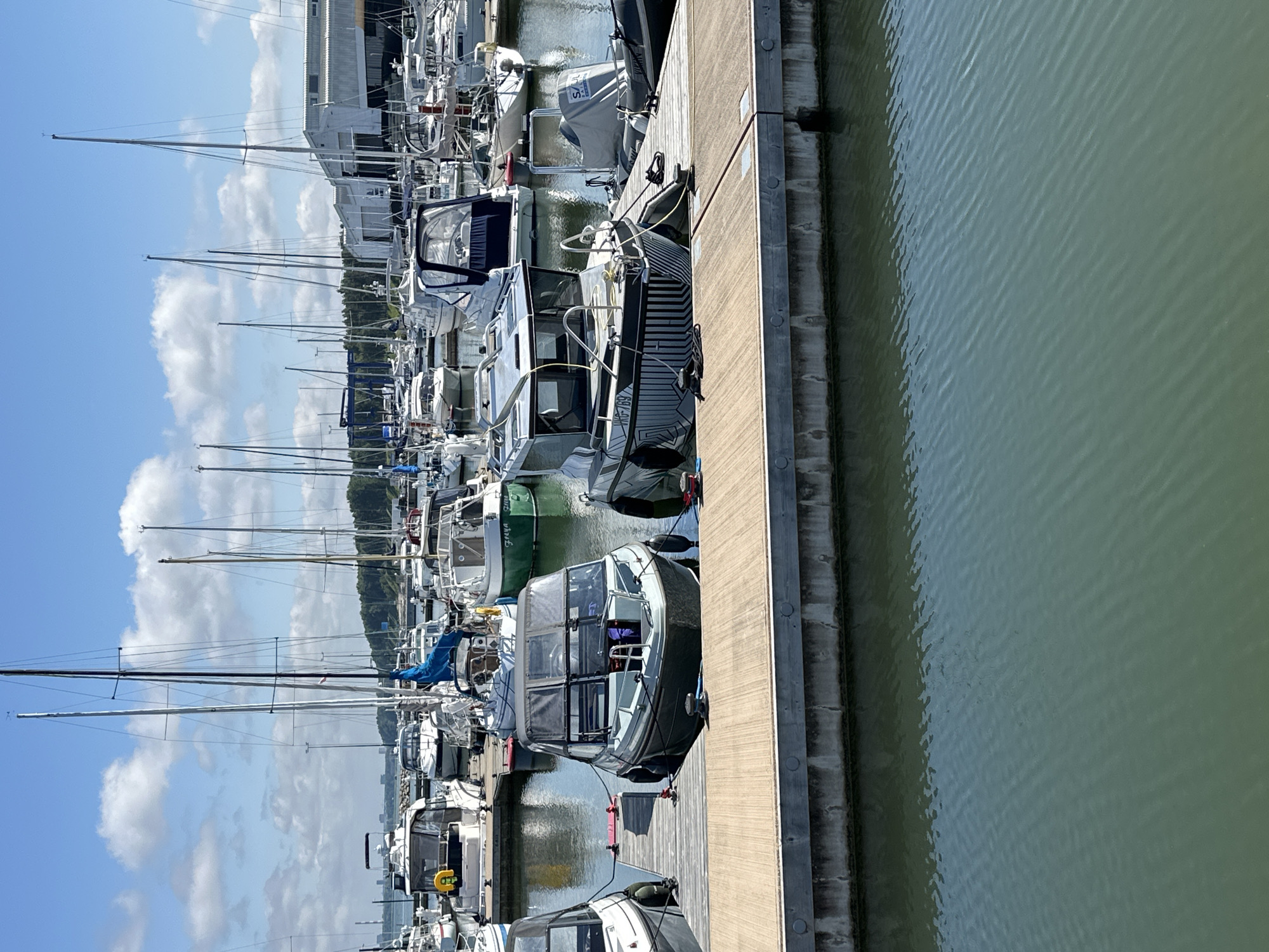 This is a full, vertical shot of a marina in Tallinn, Estonia, on a sunny day with some clouds. The foreground shows a wooden dock made of planks, with a concrete base along the water. In the water, a gray motorboat is docked to the left, and a larger, gray and black motorboat with a distinctive patterned hull is docked to the right. Several other boats, including sailboats and smaller motorboats, are visible moored in the marina further back. In the background, there are buildings, including a large industrial-looking structure, and a distant cityscape. Trees line the horizon on the right side of the frame. The water is calm and green. There are no people visible in the frame.