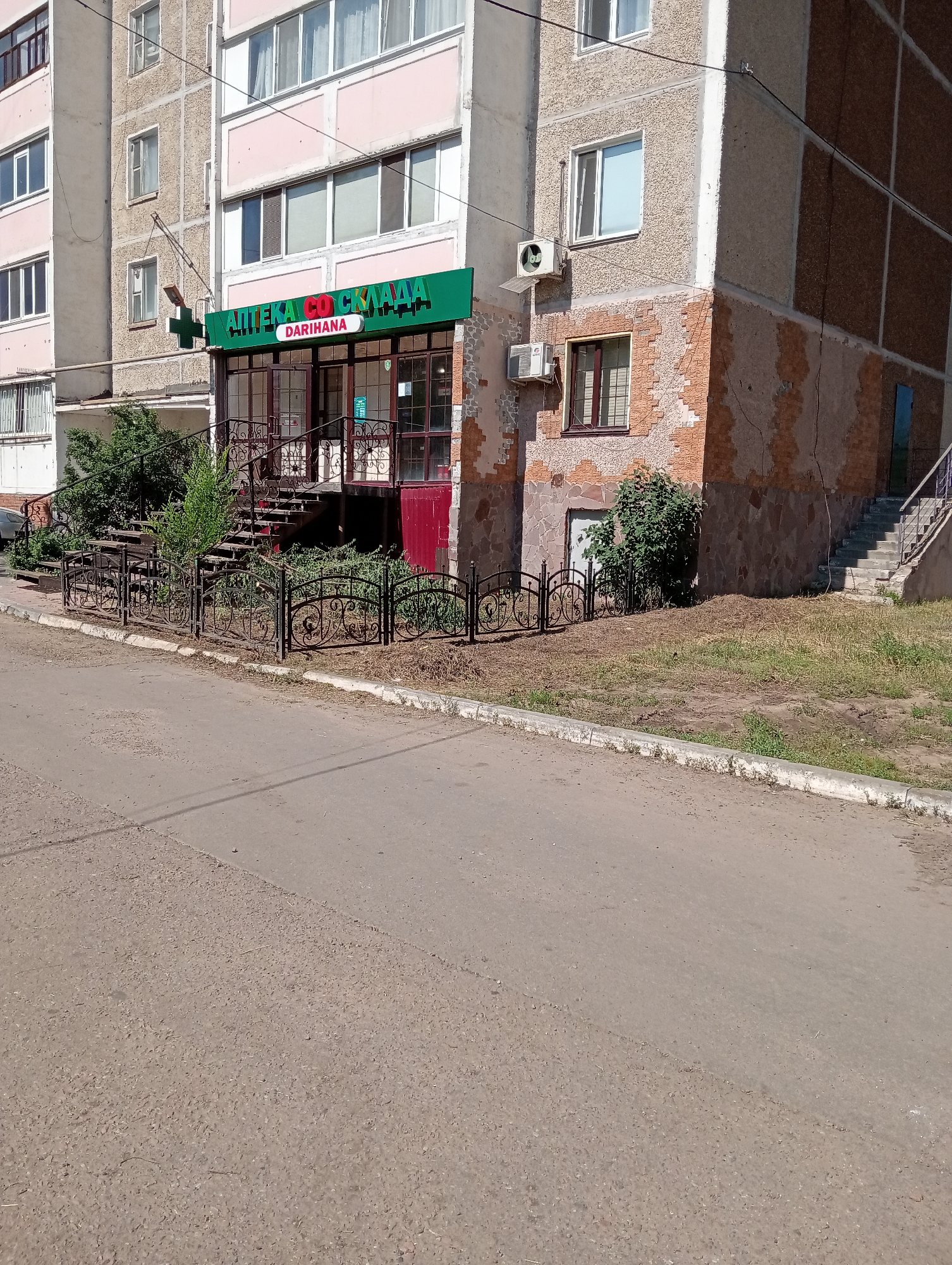 An eye-level, medium shot captures a storefront on the ground floor of a multi-story apartment building. The building's facade is a mix of rough-textured grey and brown concrete, with some areas exhibiting peeling paint and exposed brickwork. A green awning above the entrance displays text in Cyrillic script, with a smaller red label below. The entrance itself features double glass doors framed in dark metal, accessed by a set of outdoor stairs with metal railings. A small, green cross sign hangs to the left of the entrance, suspended from a metal pole. Decorative black metal fencing lines the area in front of the storefront. Adjacent to the building is a patch of dry, matted grass and several green bushes. A paved road runs along the foreground of the image, with a concrete curb separating it from the grassy area. The scene is illuminated by bright sunlight, casting shadows on the building and ground. There are no people visible in the frame.