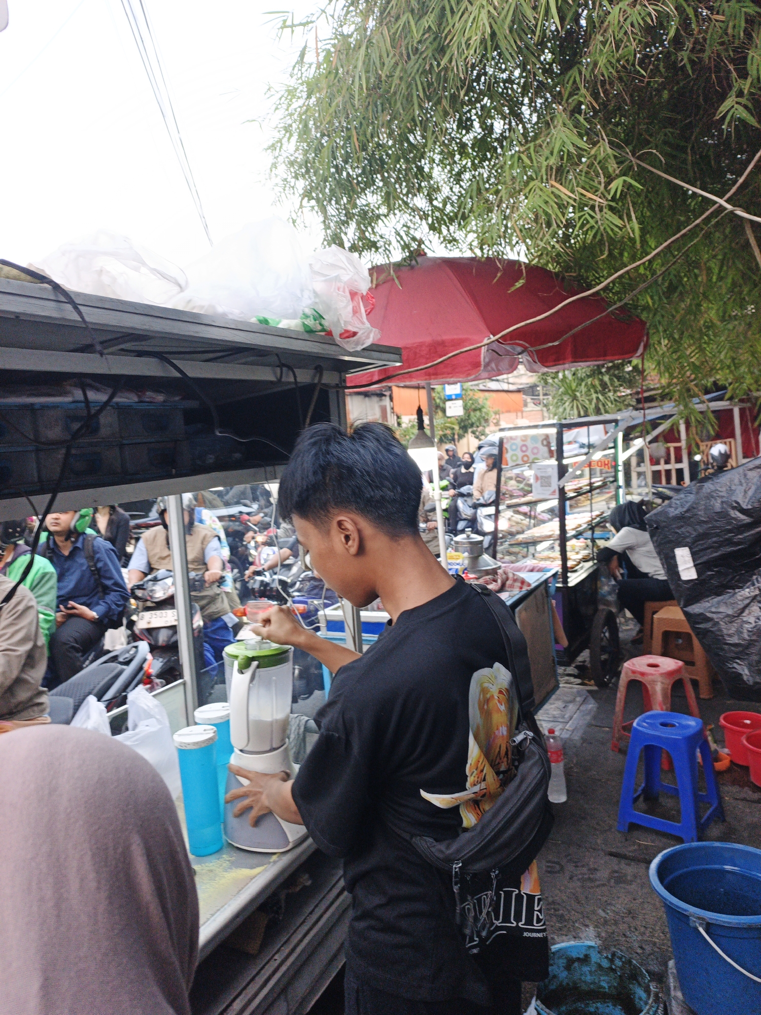 The image depicts a street scene in West Jakarta, Indonesia, likely a market or roadside vendor area. A young man in a black t-shirt with a graphic print is operating a blender, suggesting he is preparing a beverage. He is positioned in front of a stall with various items displayed, including what appear to be food items behind glass. In the background, other individuals and motorcycles are visible, indicating a bustling environment. The presence of red and blue plastic stools, buckets, and a red umbrella suggests a temporary or open-air setup. Bamboo foliage overhangs the scene, providing shade. The overall impression is that of a typical street-level commerce setting in an urban Indonesian location.