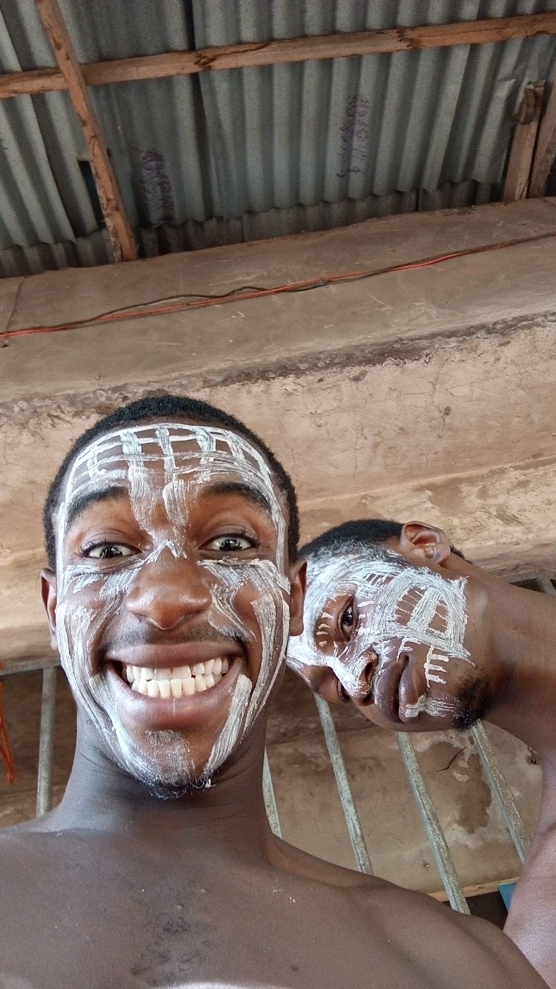 This close-up, low-angle selfie features two young men with dark complexions, both adorned with white paint in distinct patterns on their faces. The man in the foreground is prominent, smiling widely with his teeth visible, and looks directly at the camera. White paint forms a grid-like design on his forehead and is streaked across his cheeks and chin. He appears shirtless. Behind and to his right, the second man is partially visible, also displaying similar white facial paint patterns, and looks towards the camera with a more subtle expression.
The background reveals an interior or semi-enclosed space. Above the men, a corrugated metal roof is visible, supported by rough wooden beams. Below the roof structure, a concrete beam or wall runs horizontally, featuring exposed red and black electrical wires. Metal bars, possibly part of a window or security grating, are discernible in the mid-ground.
The overall mood of the image is joyful and relaxed, largely conveyed by the foreground man's wide, happy smile. The white facial paint suggests participation in a cultural event, celebration, or ritual. On one of the corrugated roof sheets, faint, dark, and indecipherable markings, possibly handwriting or symbols, are visible. The natural lighting suggests it is daytime.