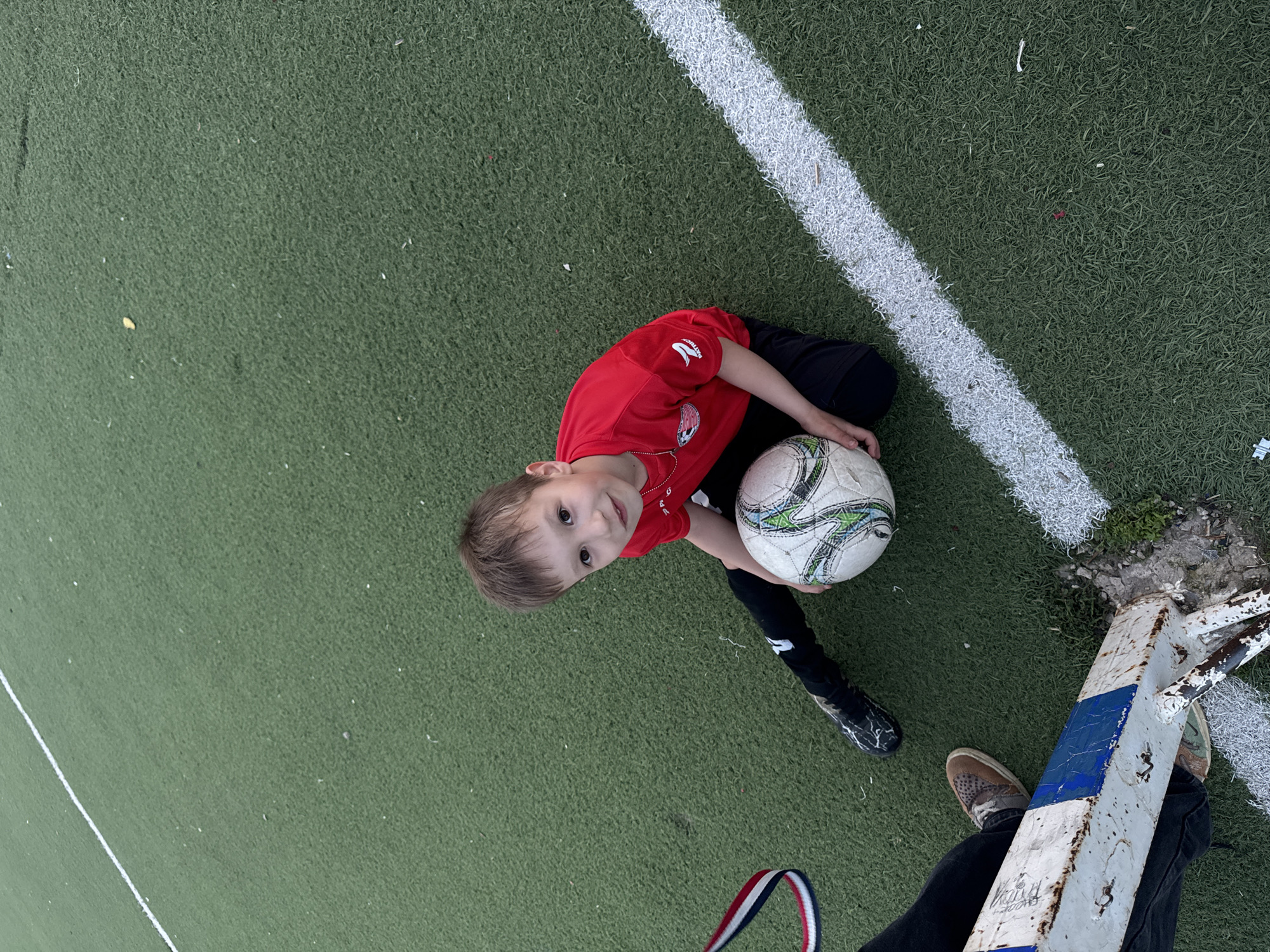 A child in a red shirt and black pants sits on an artificial green field holding a soccer ball. A white line is visible on the field. The child is positioned close to a weathered white goal post with blue accents. Another person's legs and shoes are also visible near the goal post. The picture was taken in a location with artificial grass. The city of Chaikovsky, Russia is not discernible from the image.