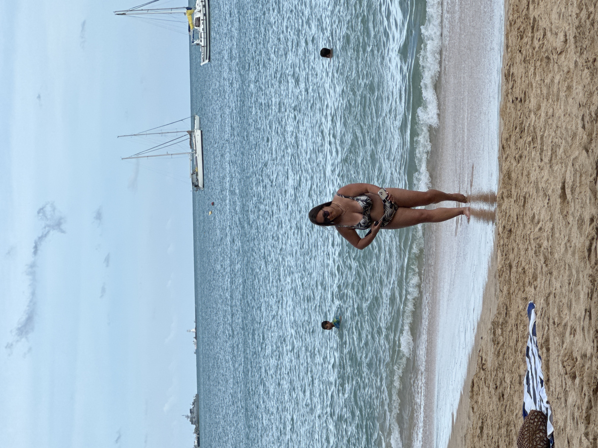 A woman in a patterned bikini walks out of the ocean onto a sandy beach under an overcast sky. She is holding a smartphone. In the water, a child is swimming, and further out, a person's head is visible. In the background, several sailboats and a larger ship are on the sea. The beach has sand and a striped towel with a straw bag on it. The location is Wichit, Thailand.