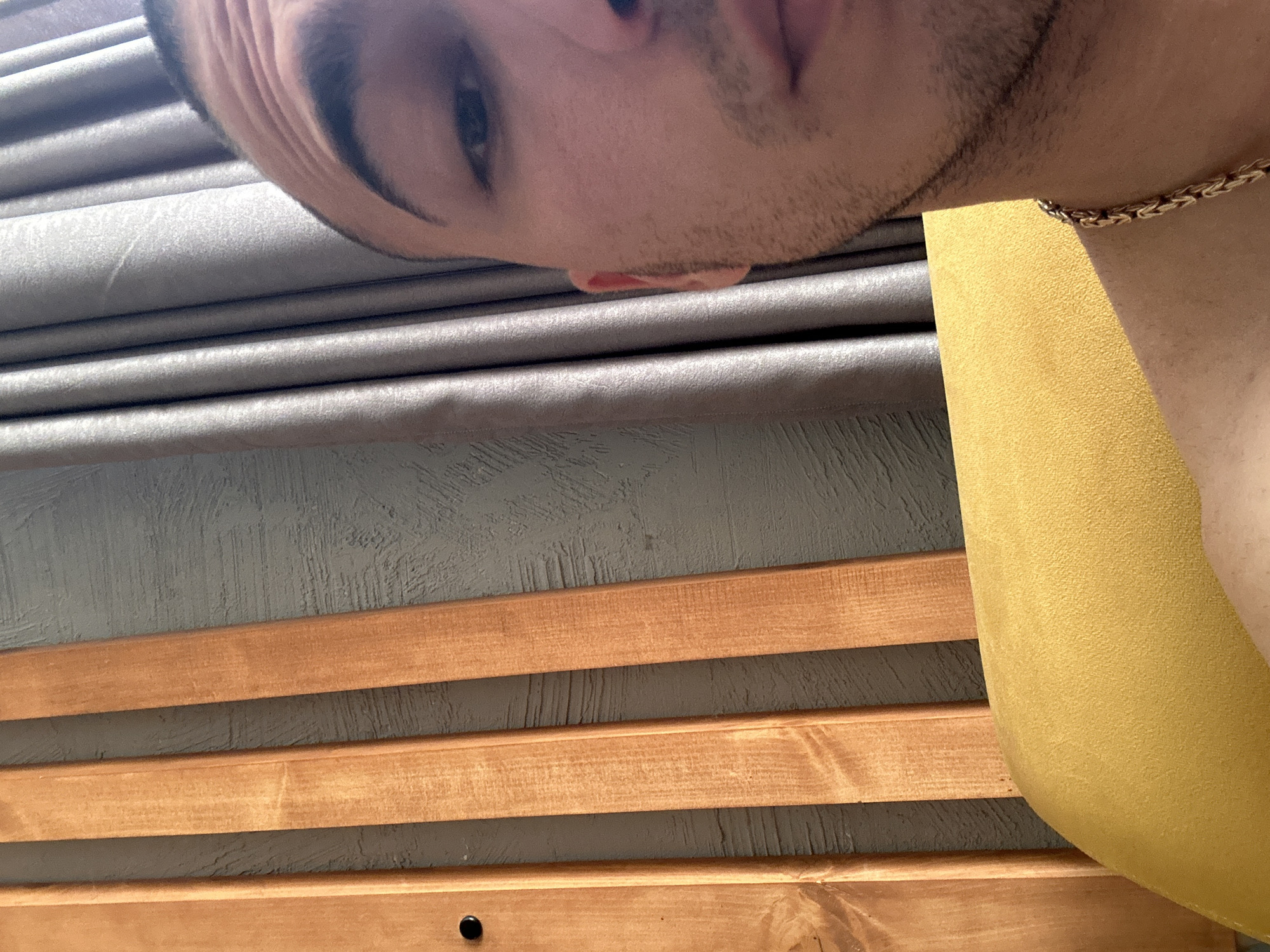 A close-up, rotated view features a person's face, neck, and shoulder against a background composed of horizontal wooden slats, gray textured wall, gray fabric, and a portion of a yellow cushion or chair. The person has dark hair and stubble, and is wearing a gold chain necklace. The wooden slats are light brown with natural wood grain patterns. The gray wall has a rough, textured surface. The gray fabric appears to be a pleated window covering. The yellow cushion or chair has a soft, textured surface. The location in the city of Unknown, Unknown is not discernible from the image.