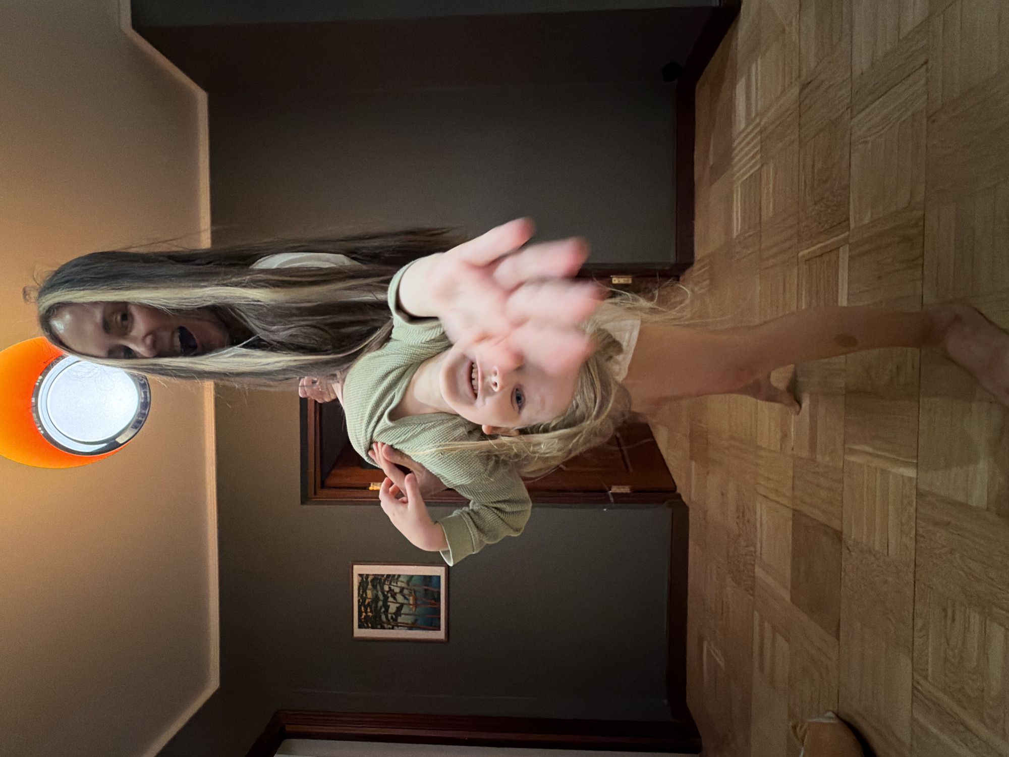 A photograph shows a light-skinned child and adult female inside a room with parquet floors, a light brown wall and a dark gray wall. The child is in the foreground with blonde hair, blue eyes, a green waffle knit long-sleeved shirt, and a hand extended towards the camera. The woman, located behind and to the left of the child, has long brown hair with blonde highlights and an open mouth. A light fixture is visible on the light brown wall. A framed picture hangs on the dark gray wall. A dark brown door with a gold-toned knob is behind the child. The scene is not explicitly identifiable as being located in Khimki, Russia.