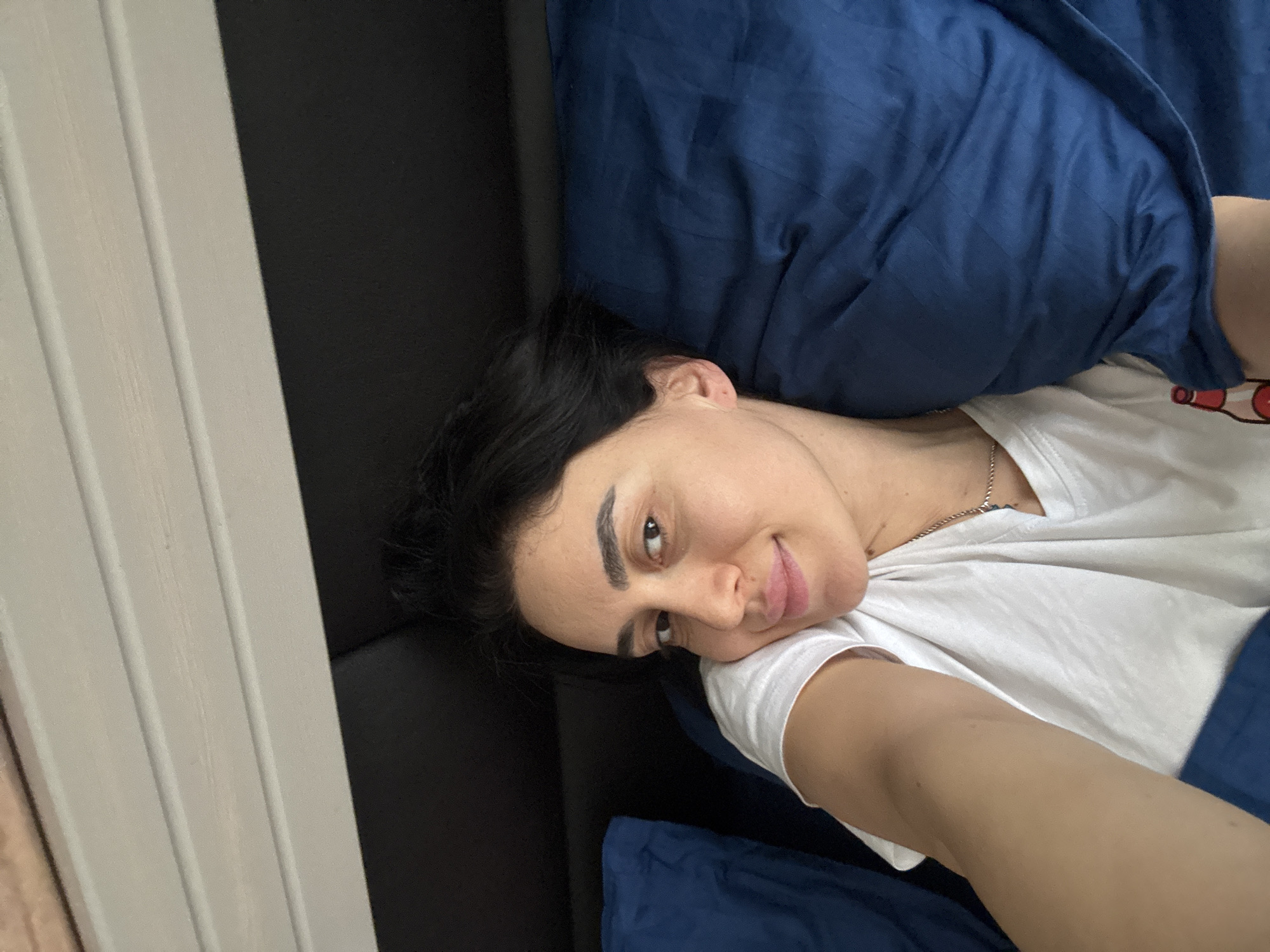 A female individual is depicted in a close-up, selfie-style photograph, lying in a bed indoors. Her head rests on a dark blue pillow, partially covered by dark blue bedding. She has dark hair, a light complexion, and dark eyebrows, and is looking directly at the camera with a subtle smile. She wears a white short-sleeved t-shirt and a silver-colored chain necklace. A dark-colored headboard is visible behind her, with a light-toned, horizontally slatted wooden paneling above it. The specific location within the city of Tbilisi, Georgia, cannot be determined from the visual evidence present in the image.