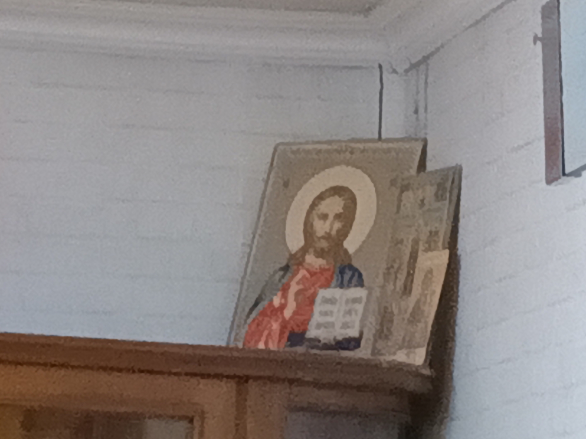 The image shows a framed religious icon of Jesus Christ, depicted with a halo and holding an open book. The icon is placed on a wooden shelf, which appears to be part of a larger piece of furniture, possibly a cabinet or a hutch. The background consists of a light-colored wall with horizontal textured lines. To the right of the Jesus icon, another framed object, possibly another icon or a picture, is partially visible. The scene suggests an indoor setting, likely a home or a place of worship. No specific location within Tatysh, Russia, can be identified from the visual information provided.